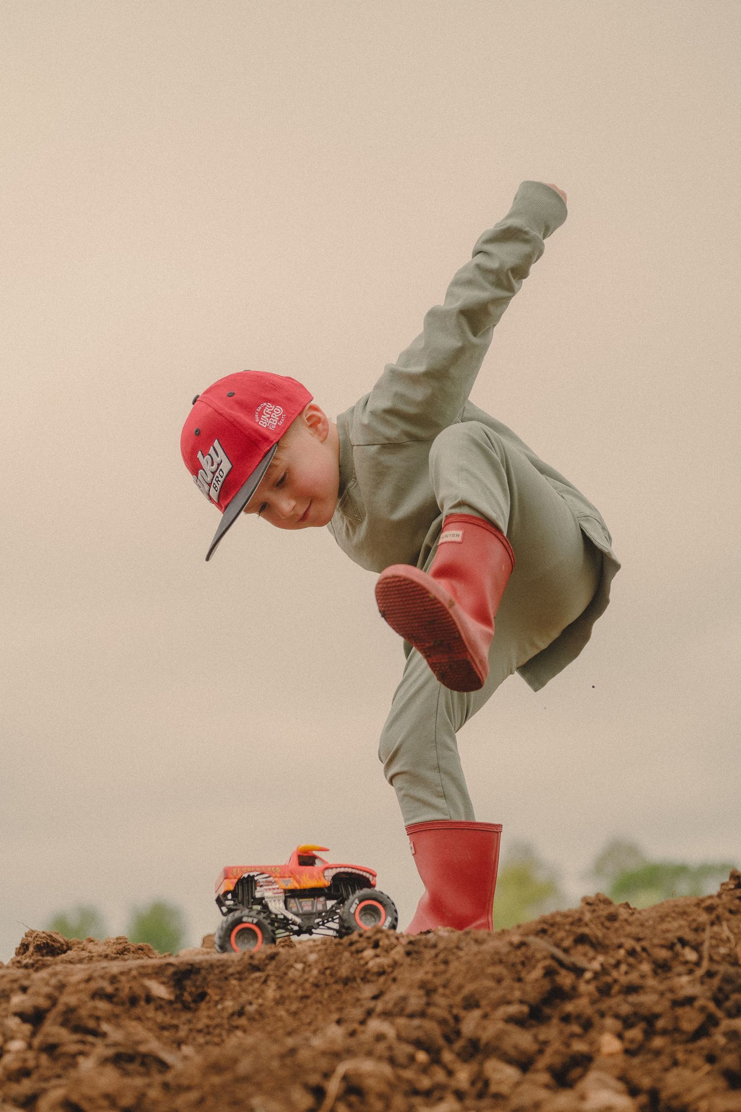 Boy in red cap and rain boots stomps near a toy monster truck on a dirt mound.