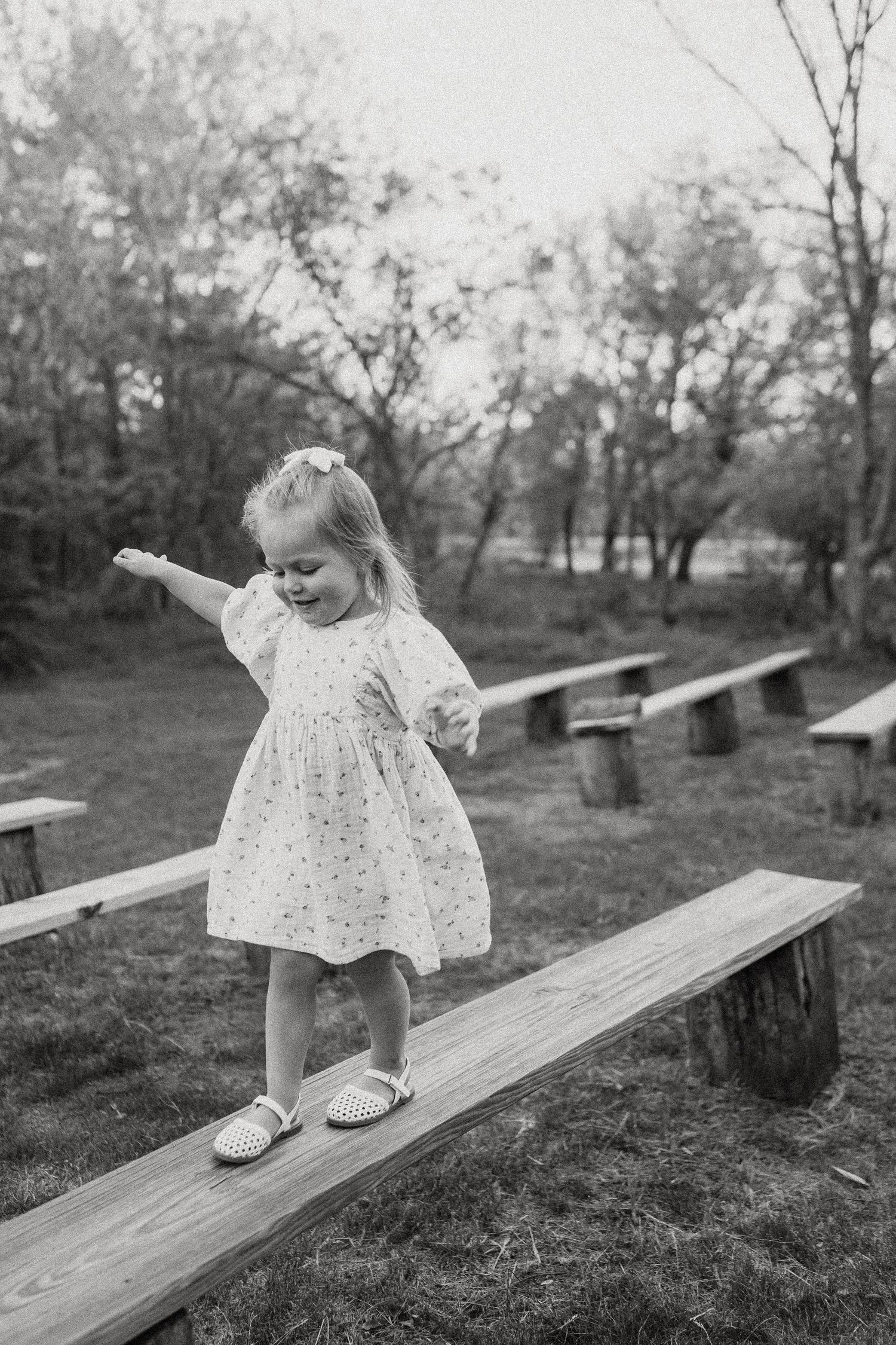 A young girl in a floral dress balances on a wooden beam in a black and white outdoor photo.