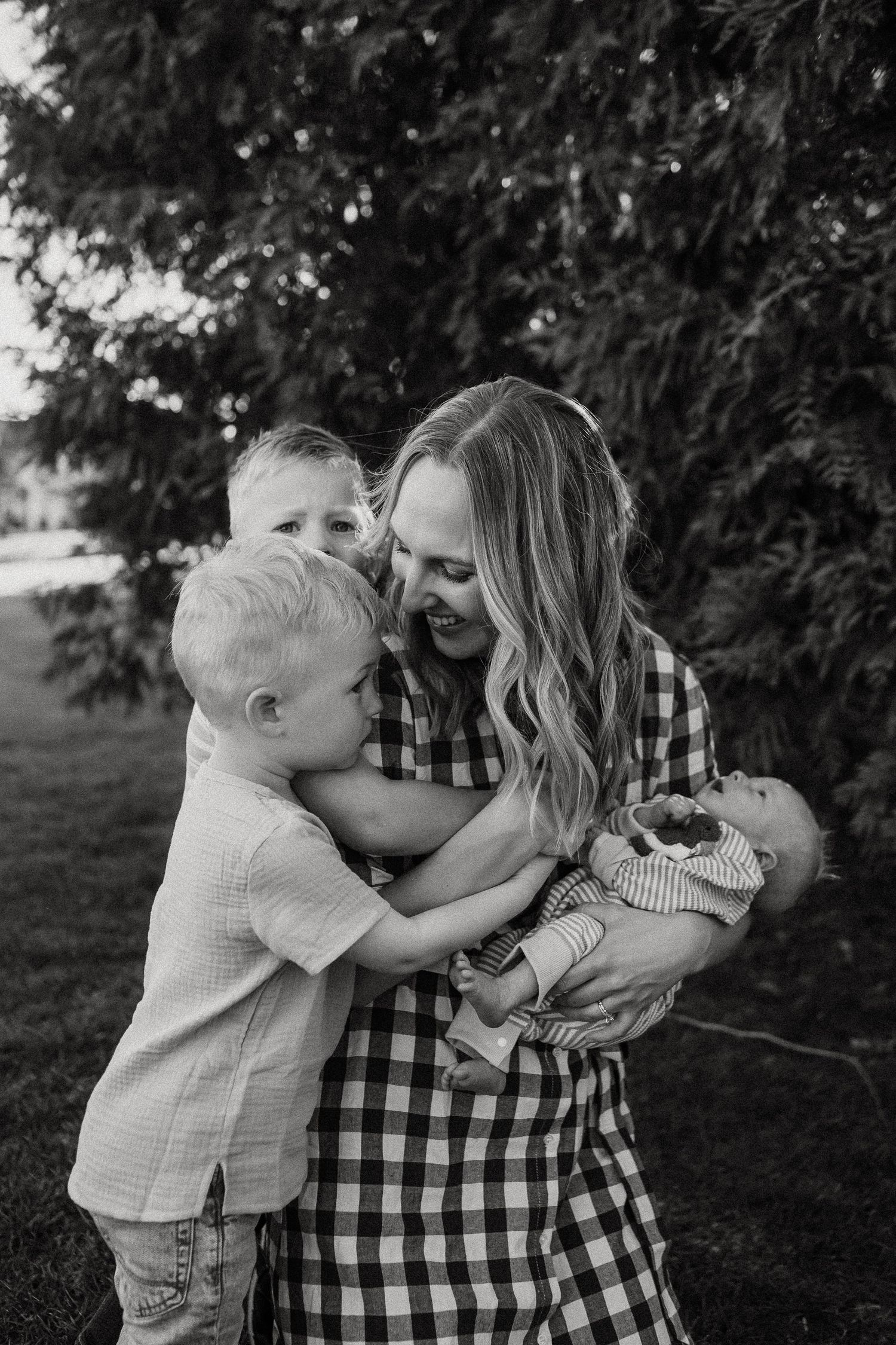 Mother in gingham dress hugging two young sons while holding newborn baby outside.