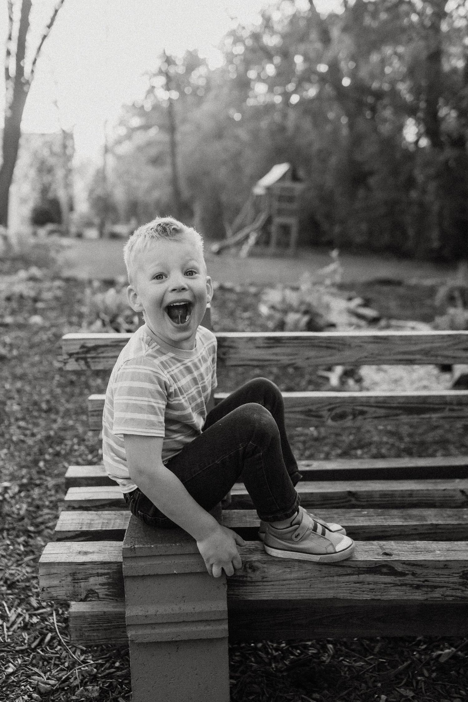 Black and white portrait of an excited laughing young boy sitting on a wooden bench outdoors.