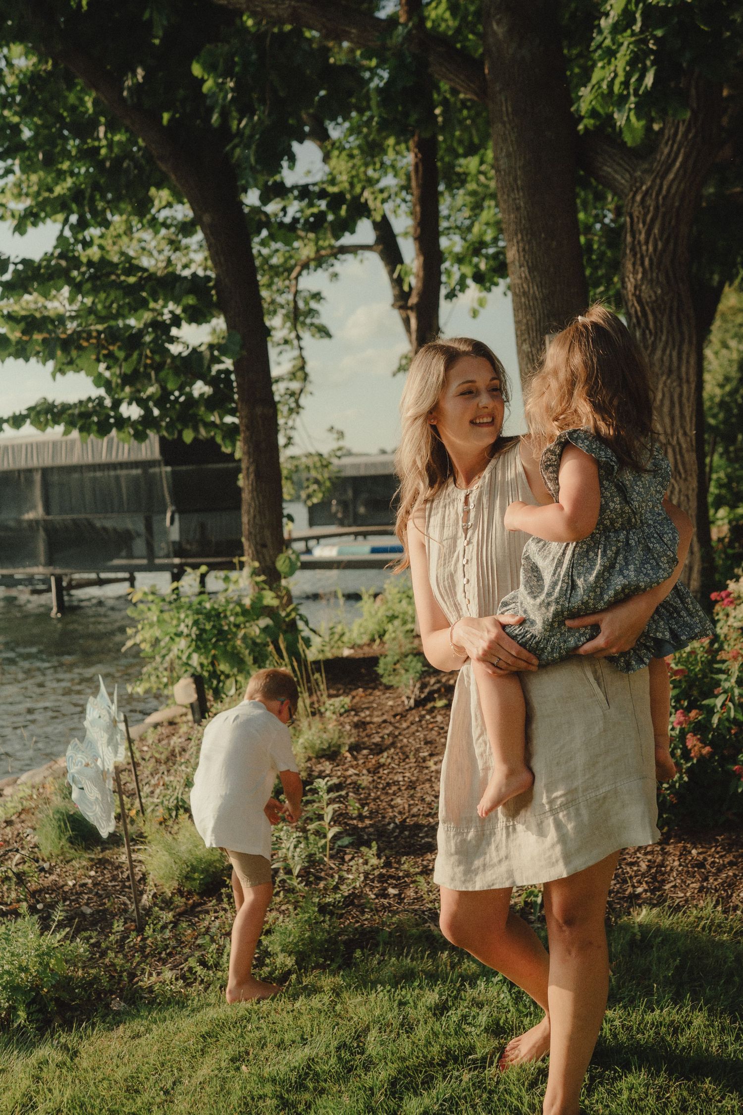 A smiling mother holds her young daughter outdoors near a lakeside garden while another child plays in the background.