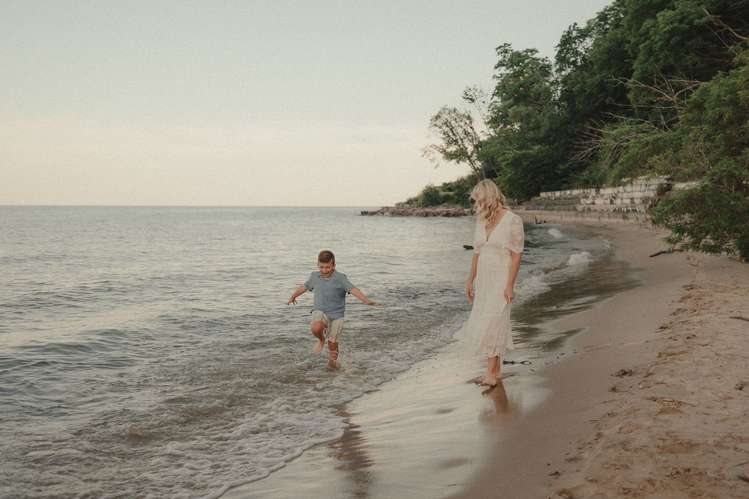 A woman in a white dress watches a child splashing in ocean waves on a sandy beach at sunset.