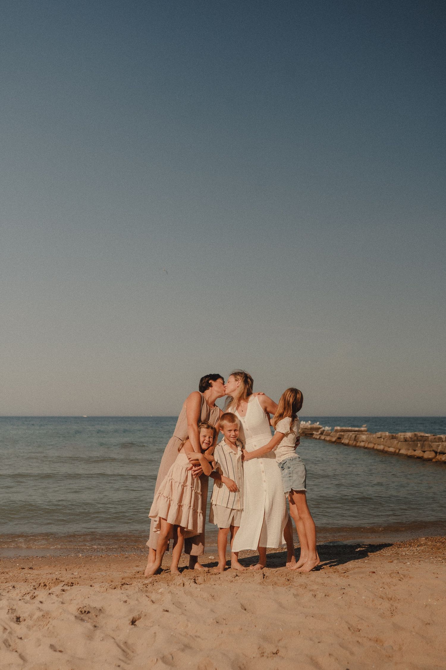 Family group photo on a sandy beach at sunset, with two women kissing a central figure while children gather around them.