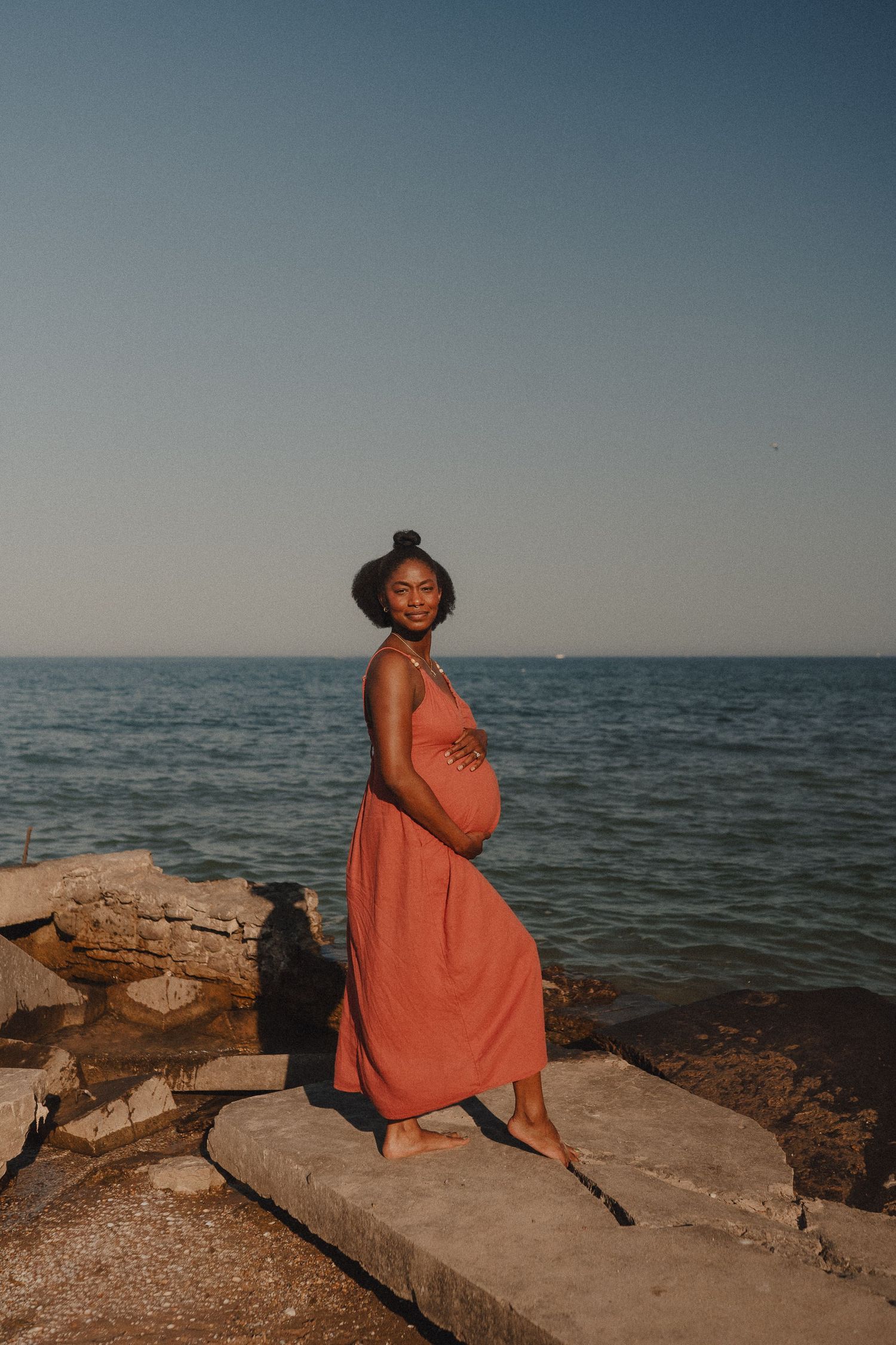Pregnant woman in coral dress standing on rocks by the ocean during golden hour.