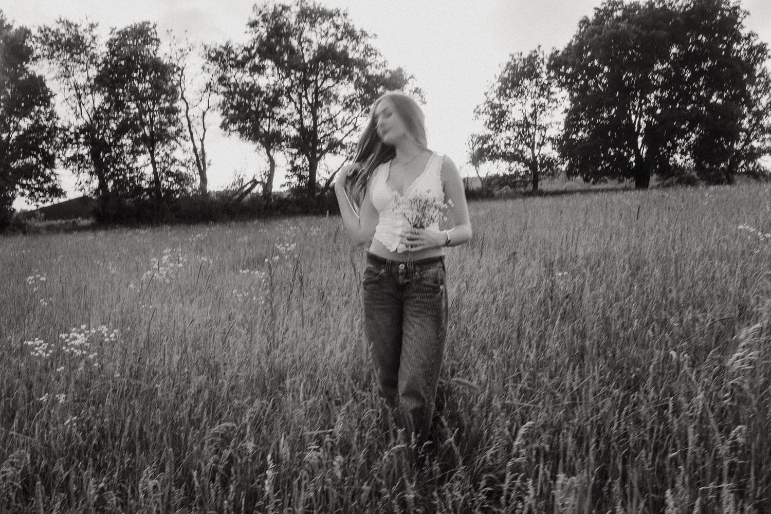 Black and white photo of a woman holding wildflowers in a lush grassy field.