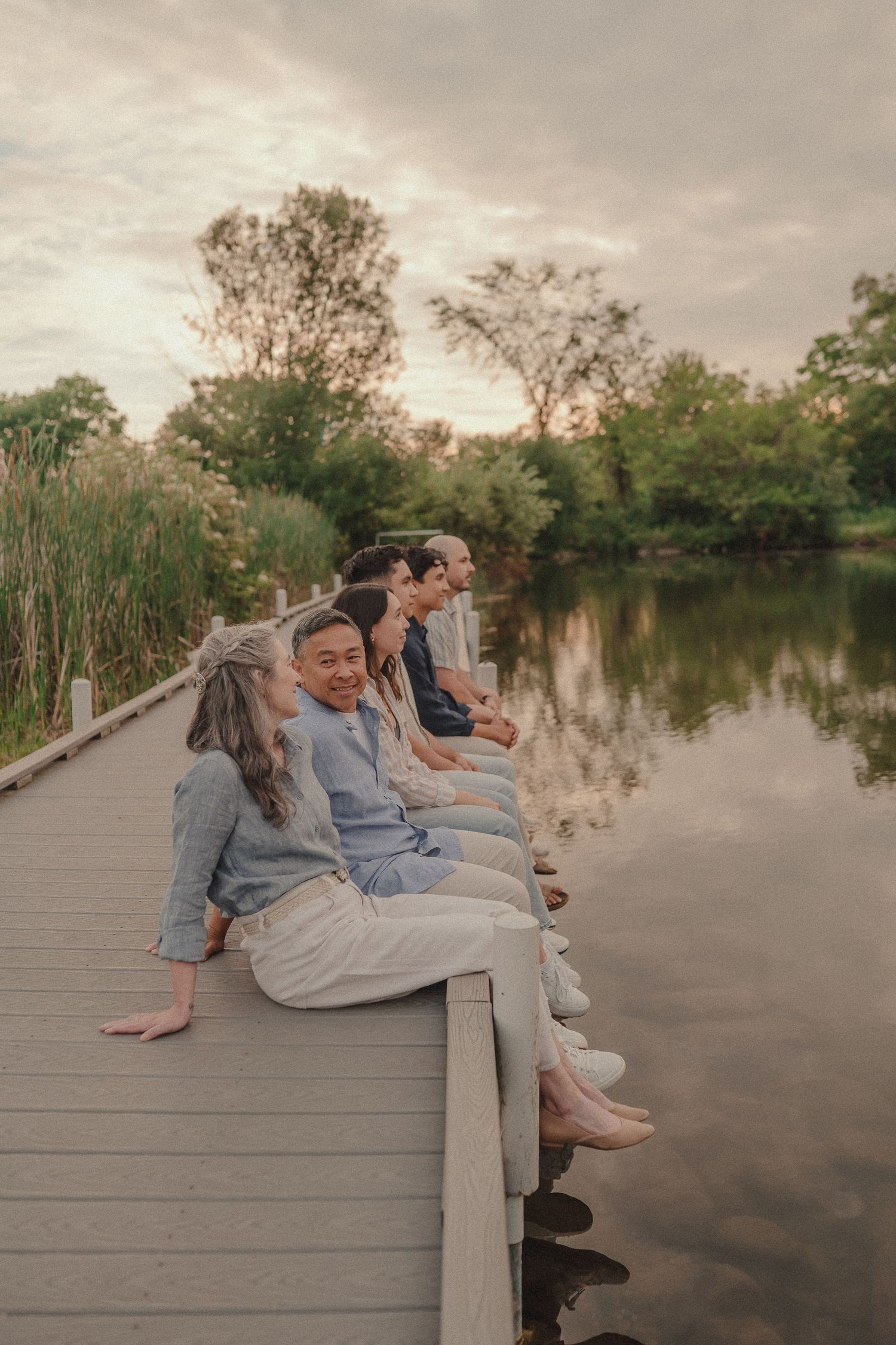 Group of friends sitting on a wooden dock by a calm lake at sunset surrounded by trees.