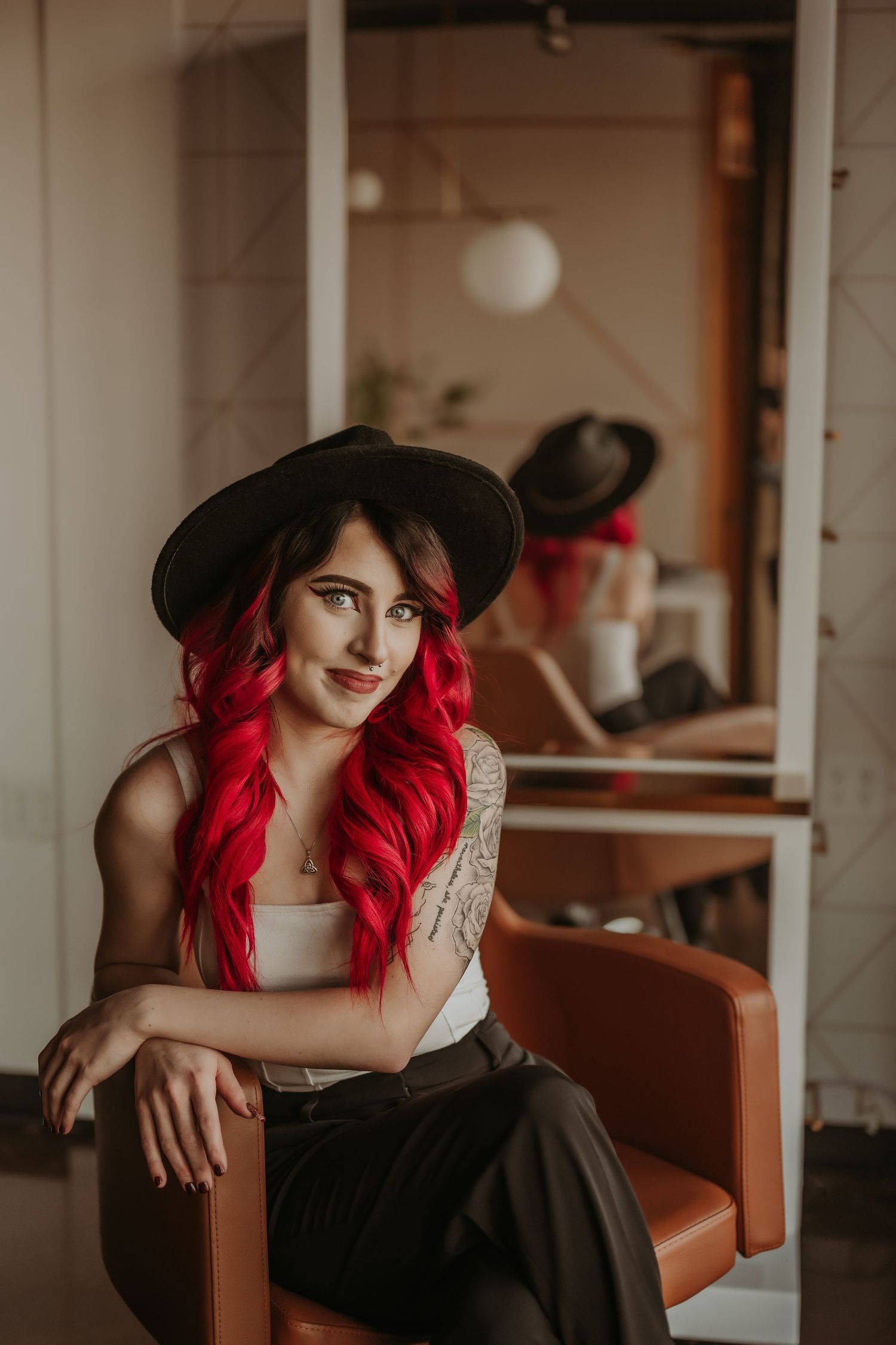 Portrait with black hat and vibrant hair seated in orange salon chair with mirror reflection.