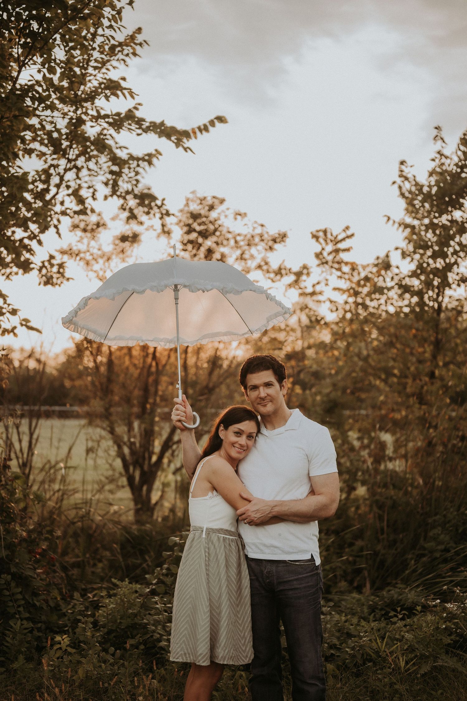 A couple embraces under a white umbrella during golden hour in a natural setting.