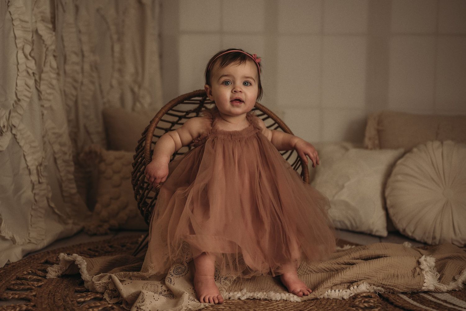 Baby in flowing pink dress sits against wicker chair in vintage-style portrait.