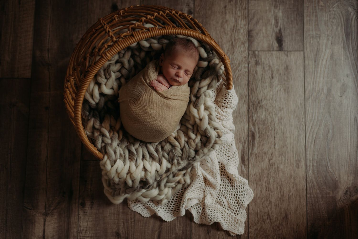 Sleeping newborn baby nestled in chunky knit blanket inside woven basket on wooden floor.
