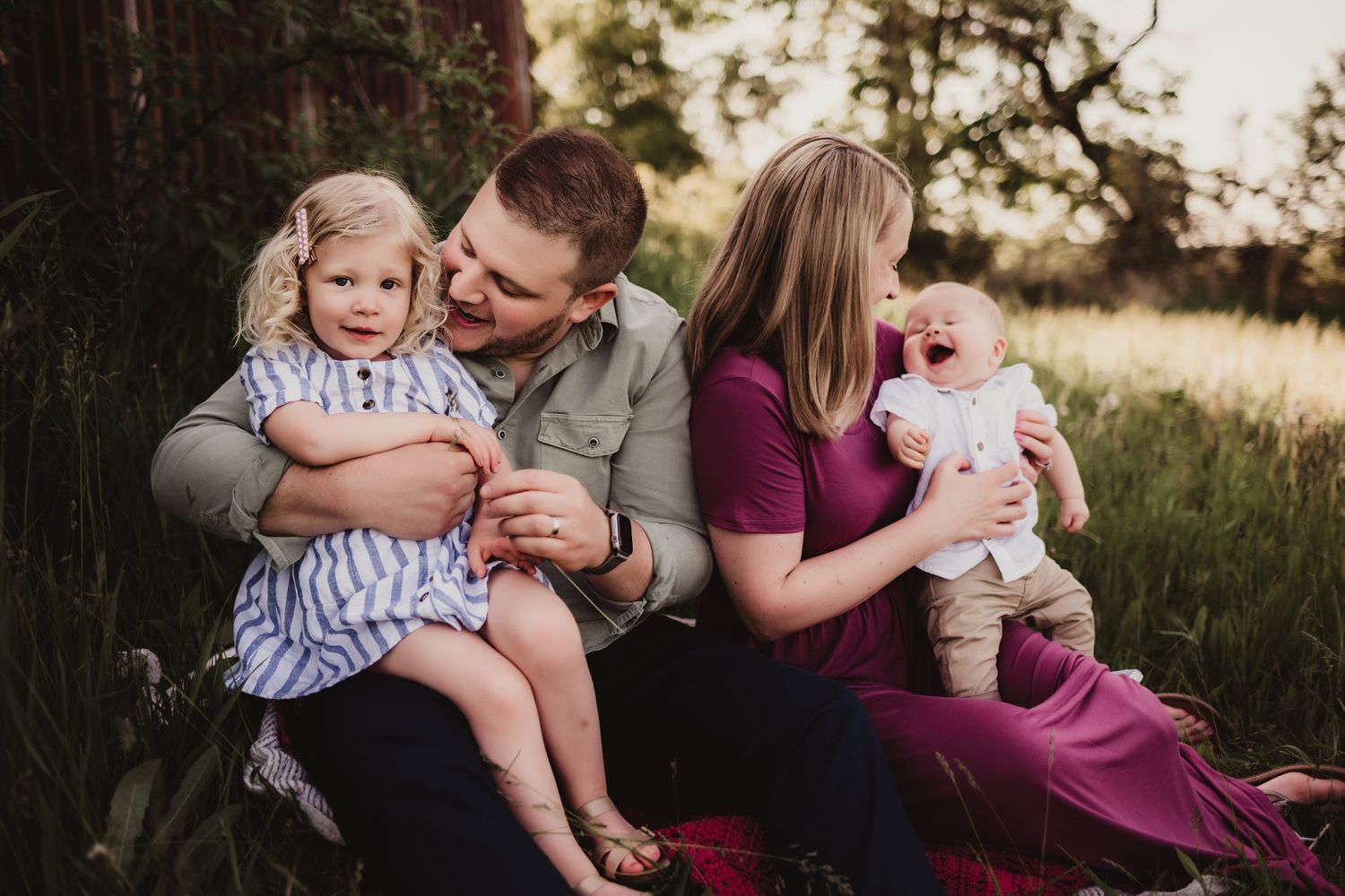 A family sits together on a blanket near a weathered barn during a sunset photo session.