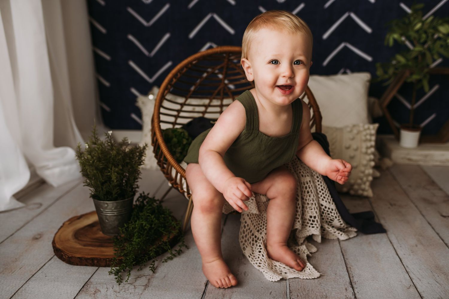 A small child in olive romper sits near potted plants and wicker furniture.