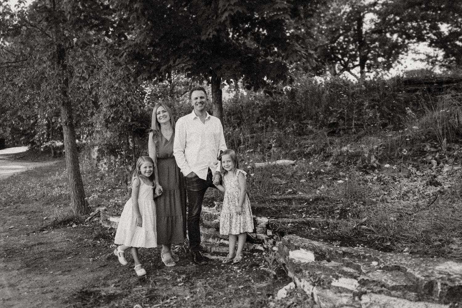 A black and white portrait of a family standing together in a wooded outdoor setting.