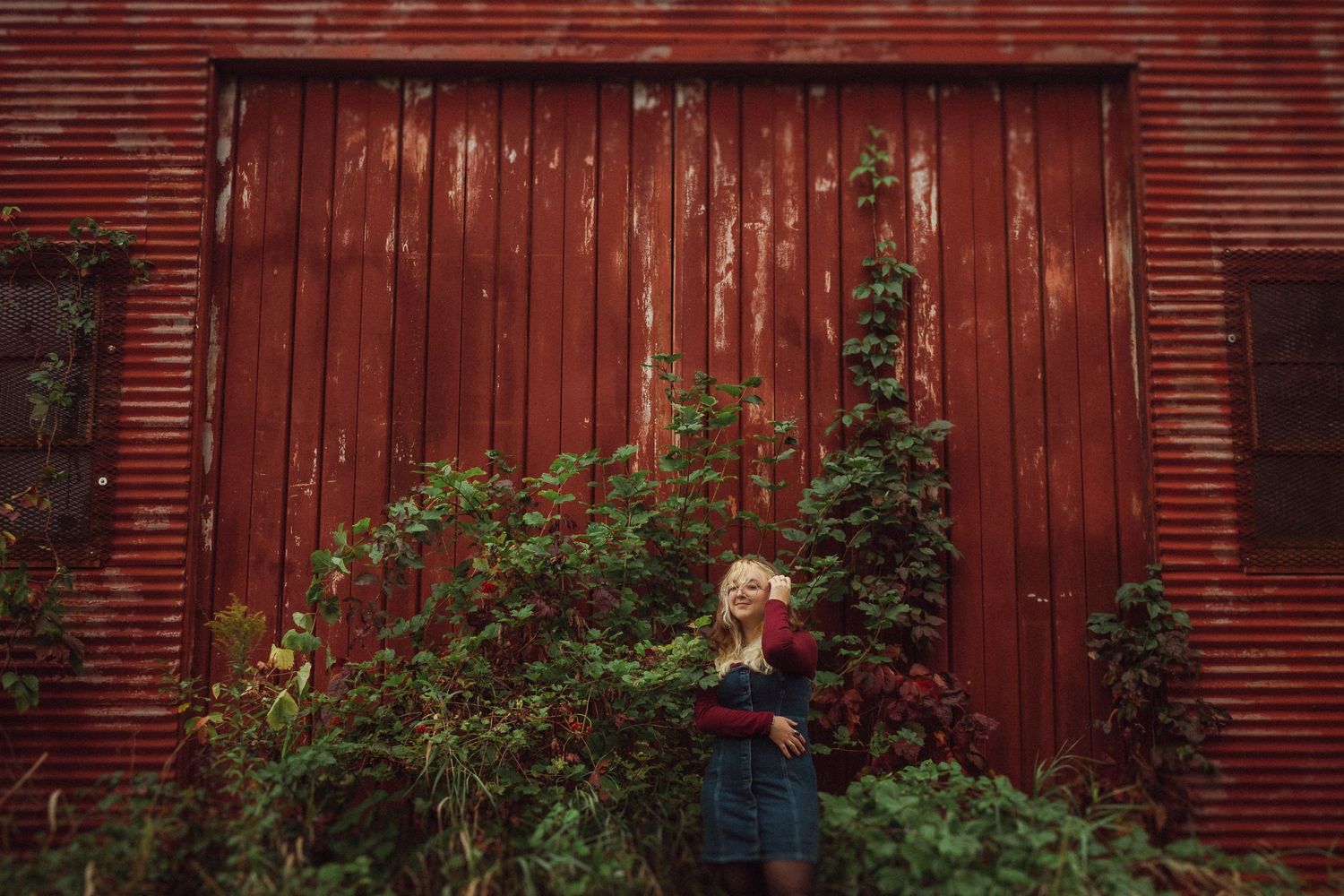 Figure in denim dress and sweater stands against rustic red barn with climbing vines in moody outdoor setting.