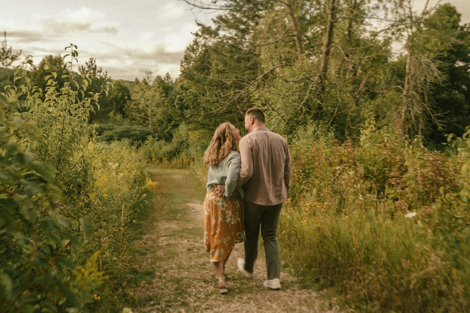 A couple walks together down a nature path surrounded by lush greenery on a cloudy day.