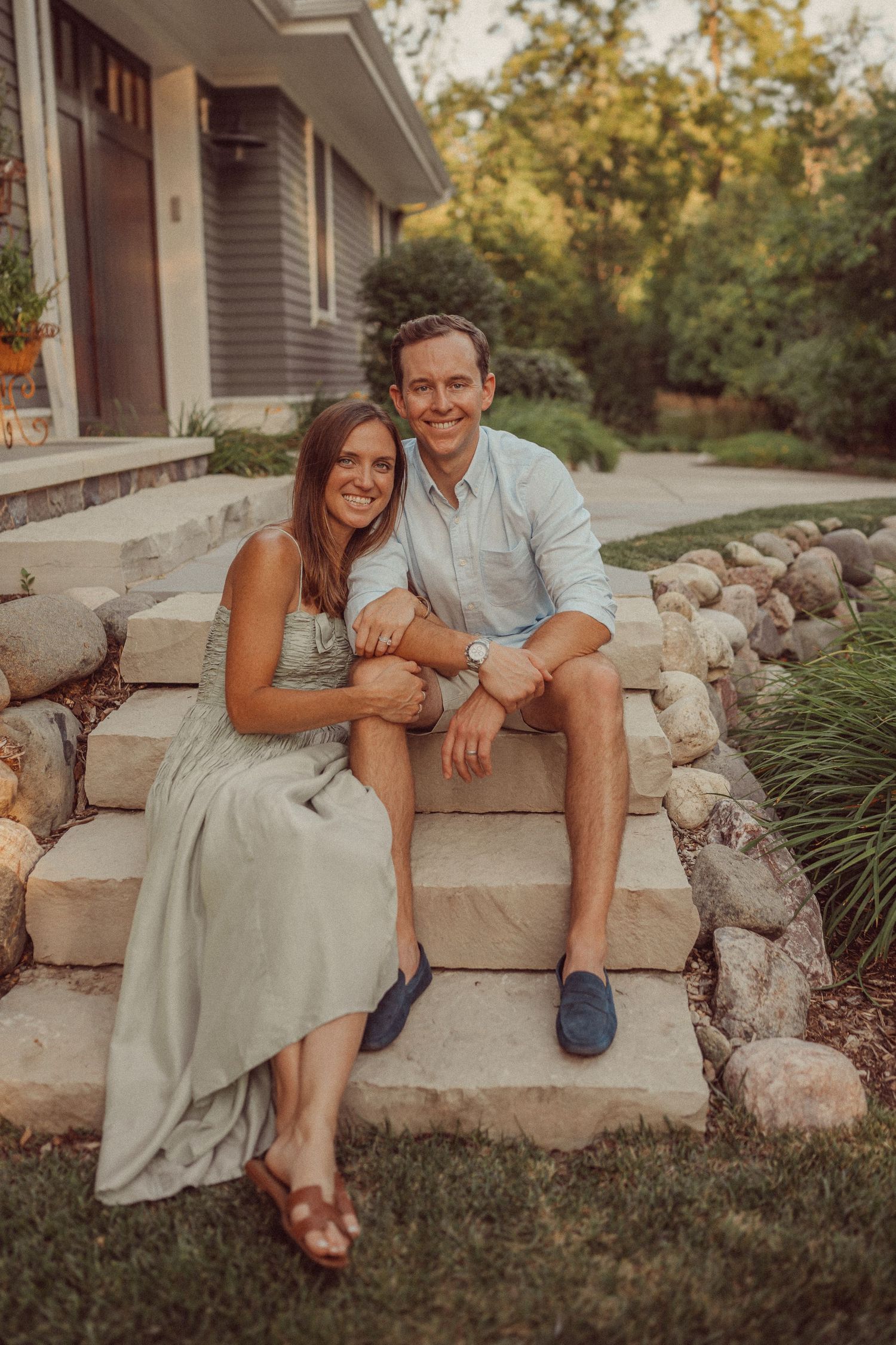 Couple sitting together on stone steps outside a gray house.