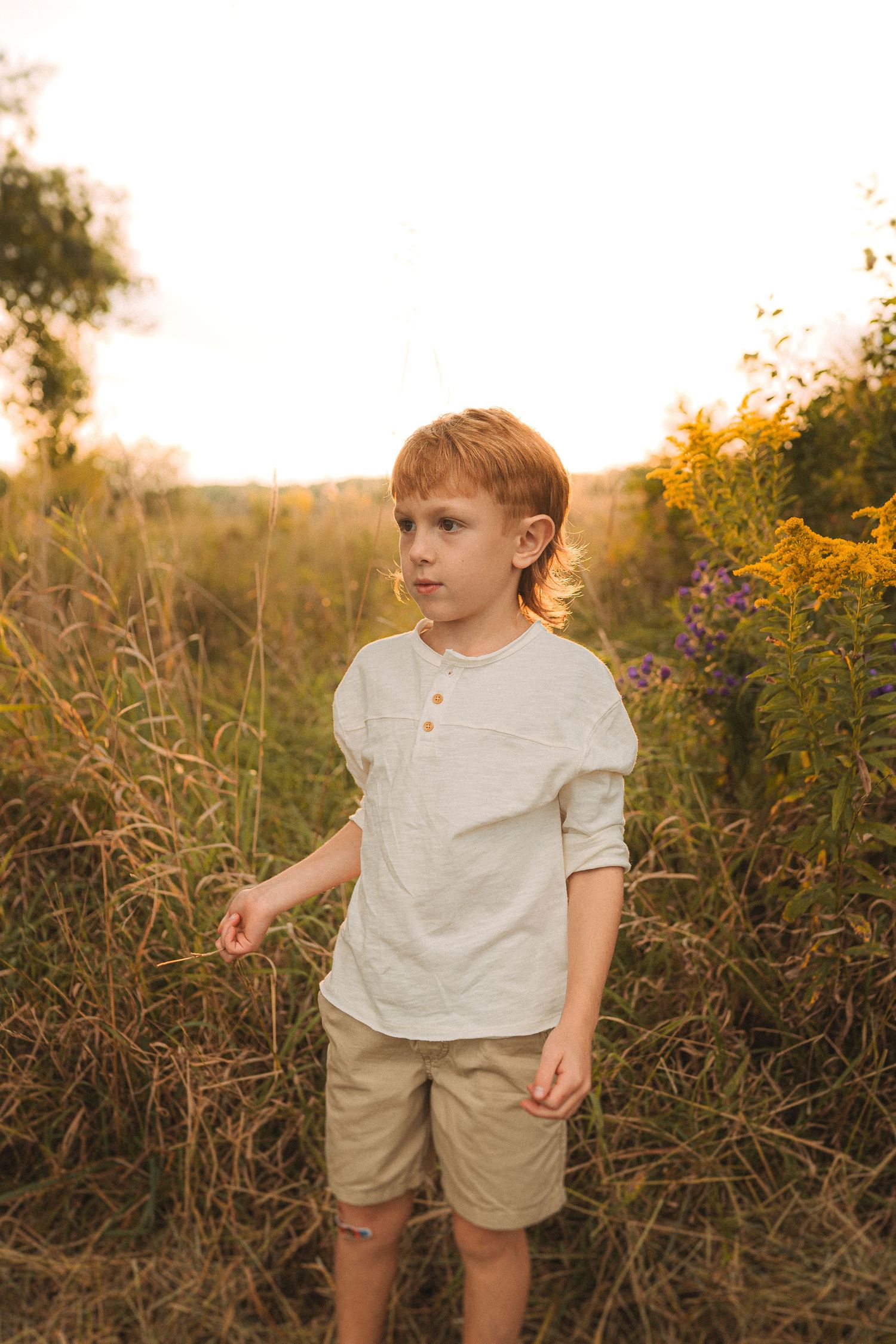 Child in white shirt and khaki shorts standing in tall grass at sunset.