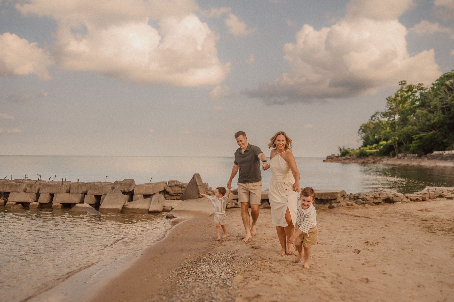 Couple walking their dog along a scenic tropical beach with rock breakers and cloudy sunset sky in background.