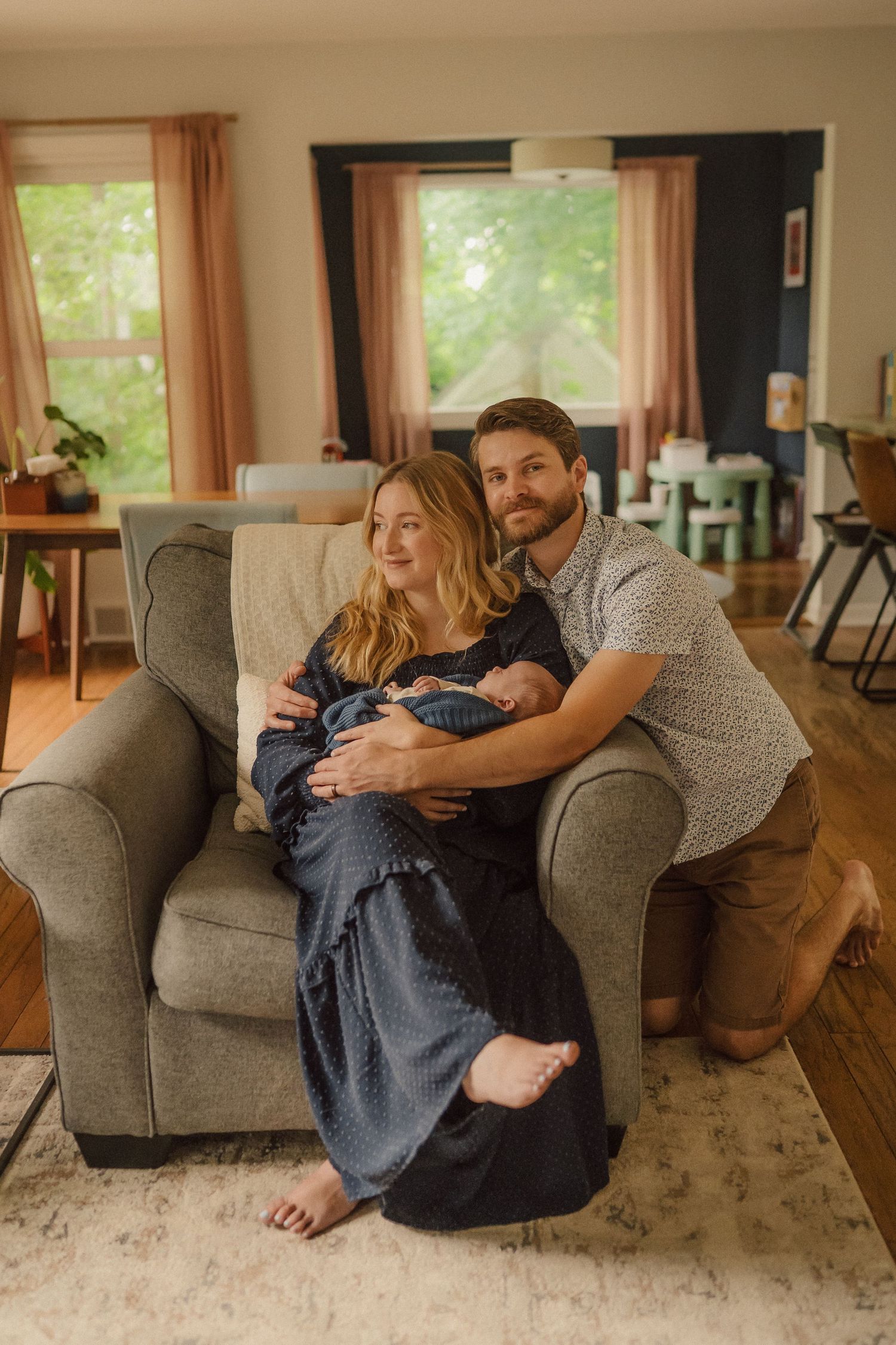 Couple cuddling together with infant on grey armchair in sunlit living room with warm decor.