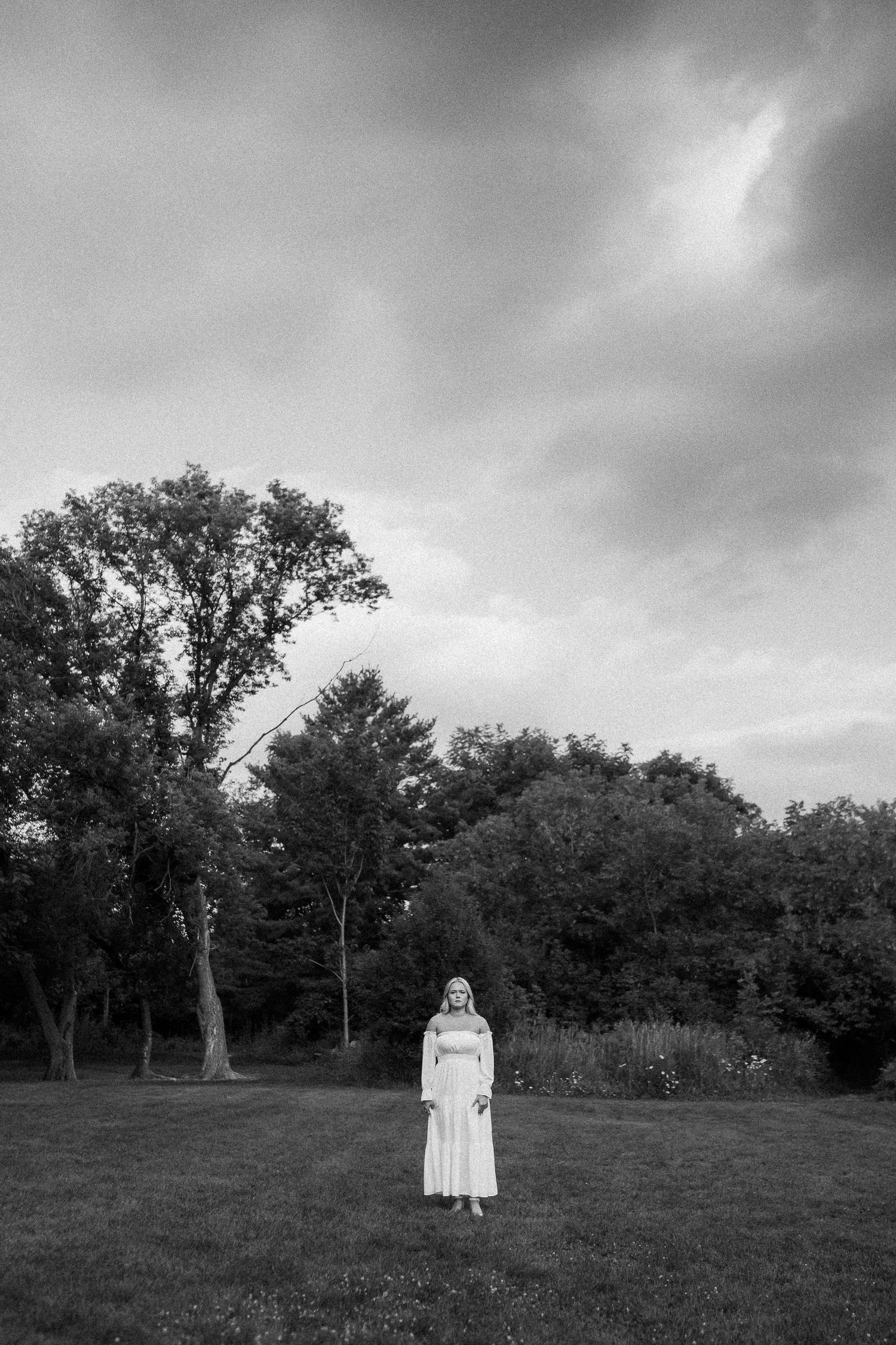 A lone figure in a white dress stands in a grassy field under moody clouds in black and white.