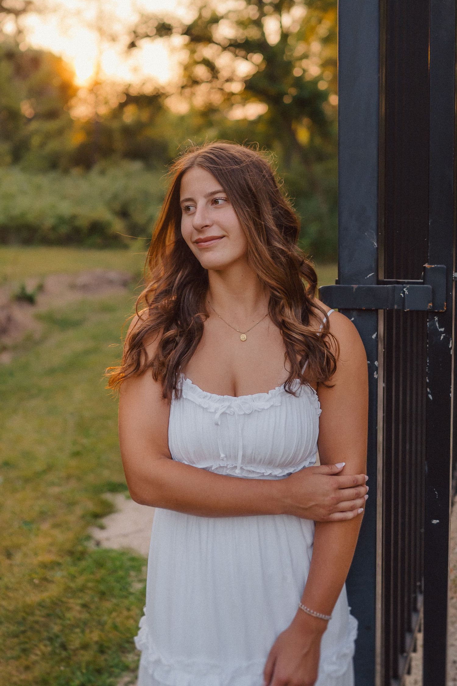 A person in white summer dress poses by dark post during sunset.