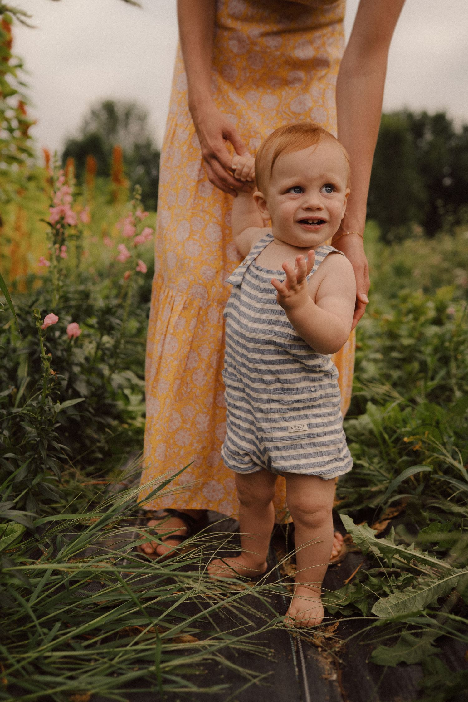 A small child in a striped romper stands in a garden while being supported by someone in a yellow dress.