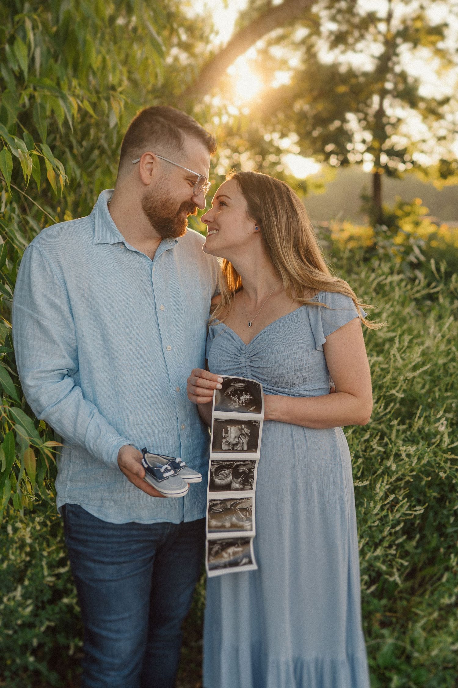 Couple shares intimate moment while holding ultrasound photos in a sunlit outdoor setting.