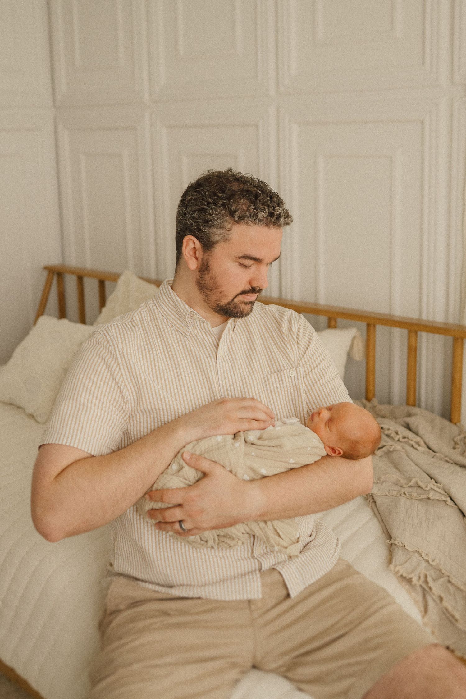 A parent in a striped shirt holds a sleeping newborn on a bed with white bedding and wooden headboard.