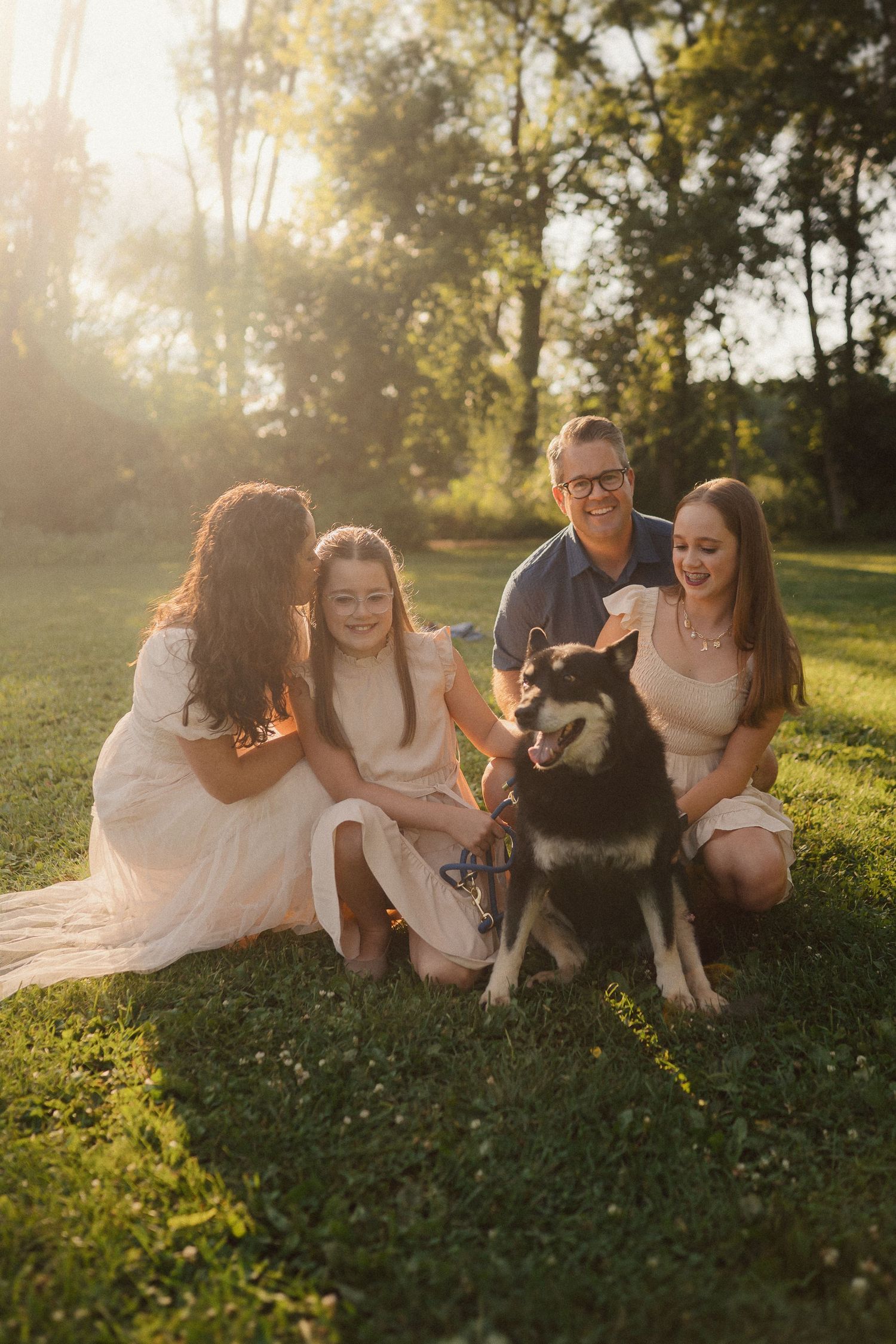 Family sitting on grass with their husky dog during golden hour sunset.