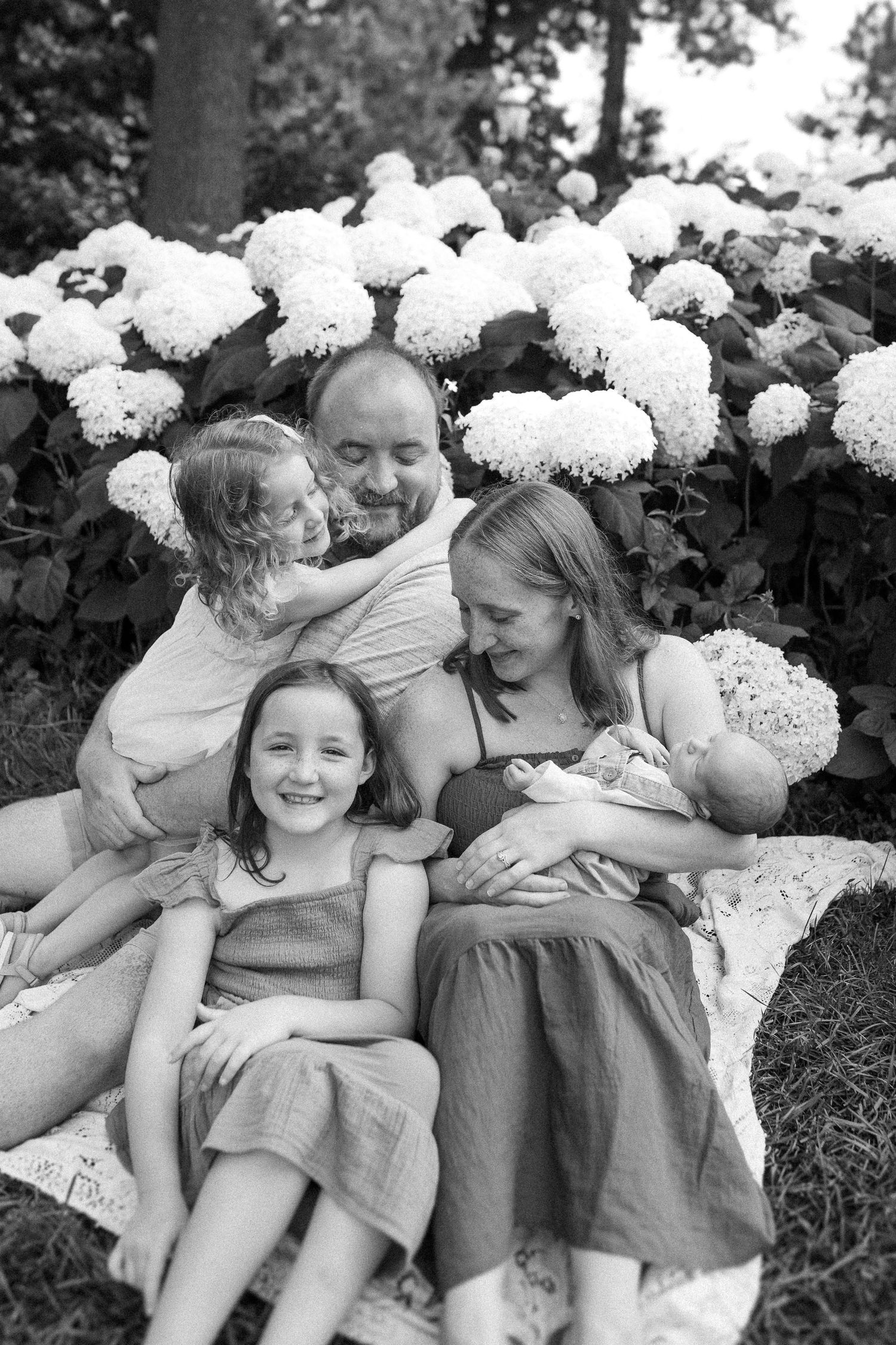 Black and white portrait of three people laying in front of blooming white hydrangea bushes in a garden.