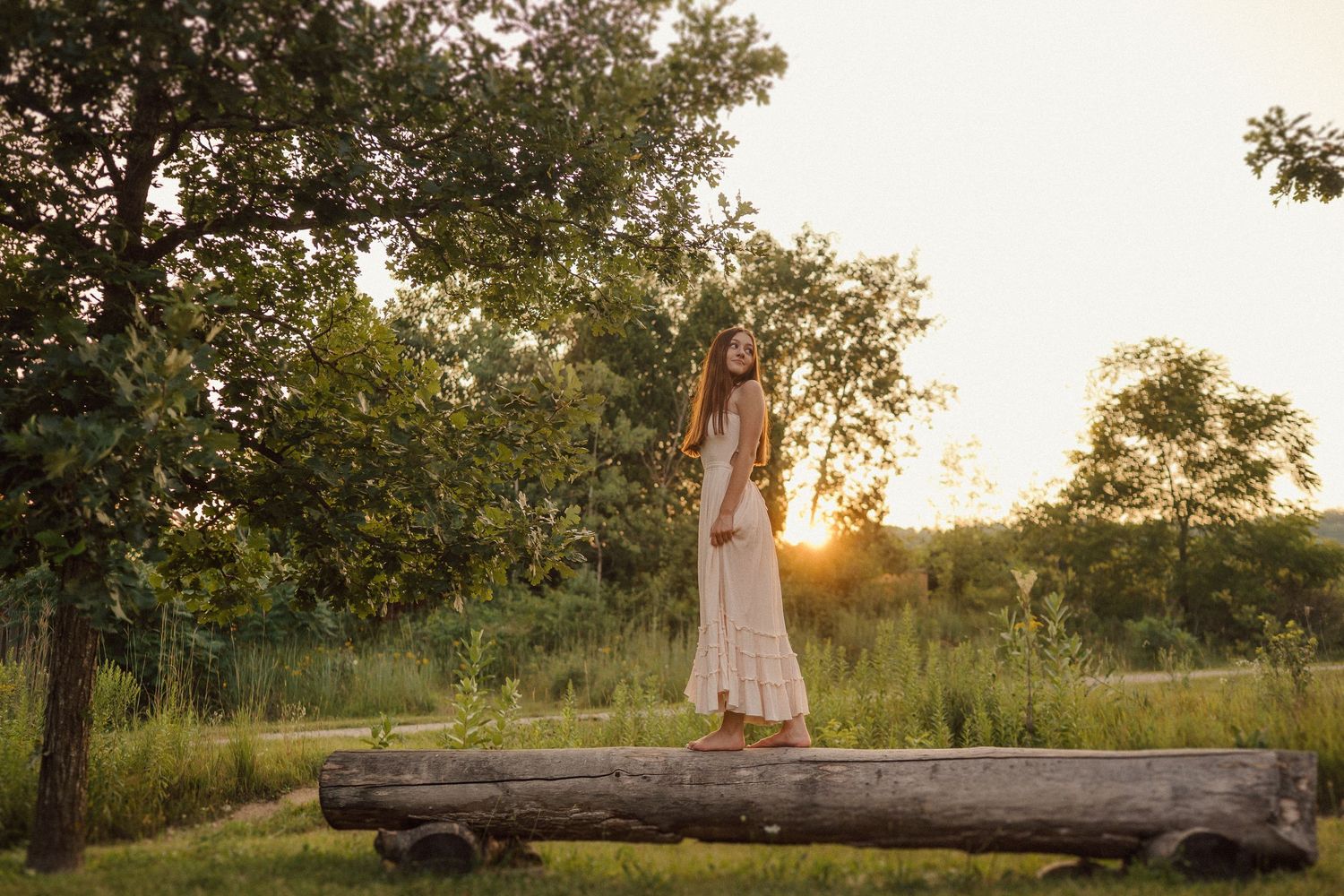 Figure in a flowing dress stands gracefully on fallen log during golden hour in nature setting.