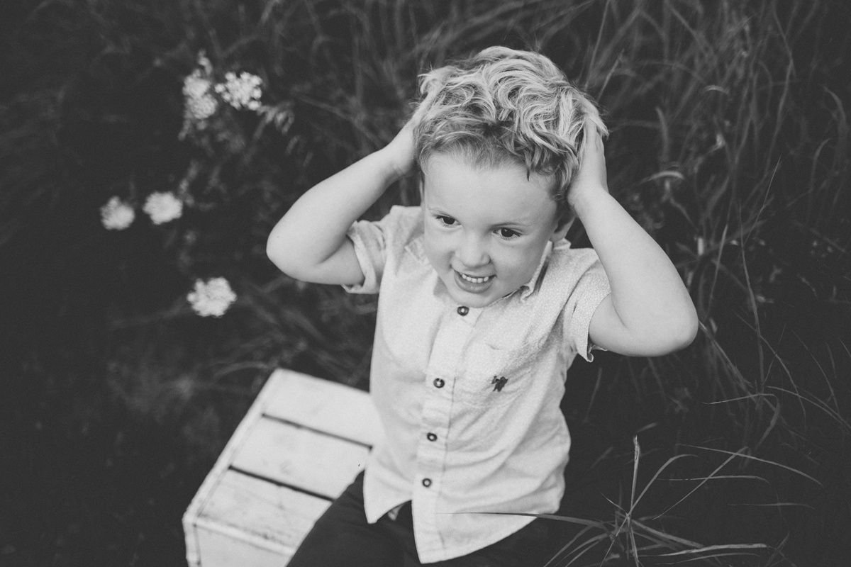 A black and white portrait of a playful child grabbing their hair while sitting next to white daisies.
