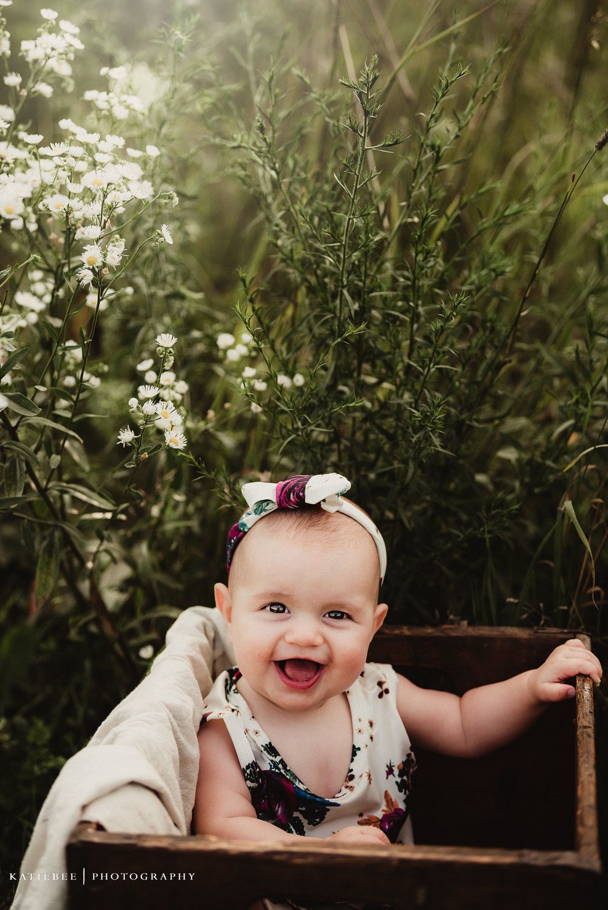 Joyful baby in floral outfit and bow laughs while sitting in wooden box outdoors.