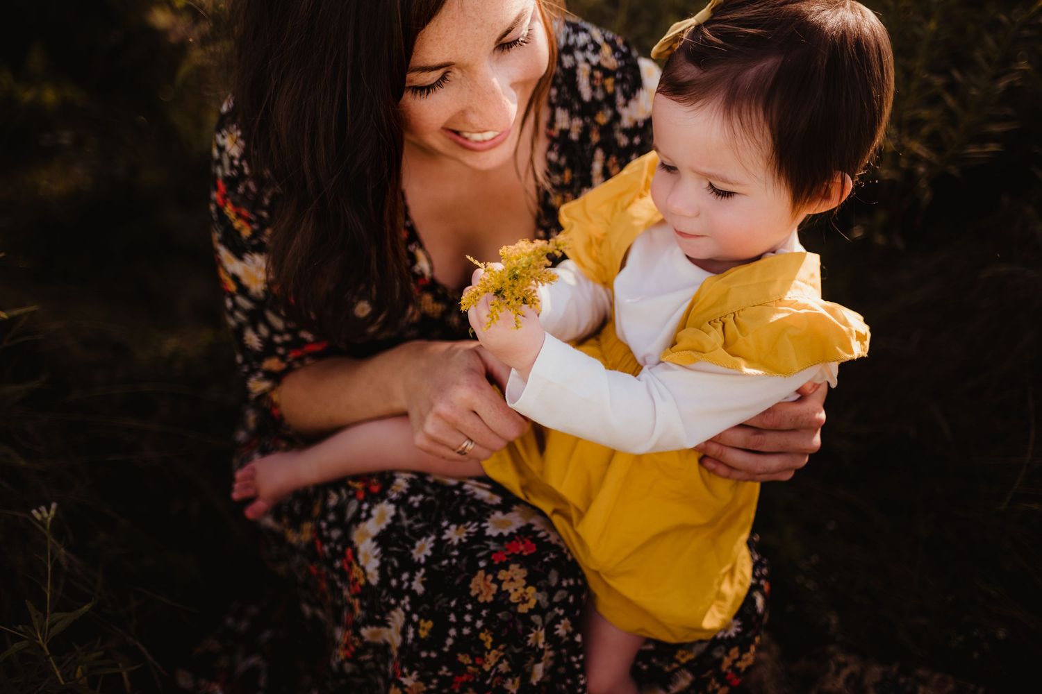 Parent and child in floral dress and yellow outfit share tender moment outdoors.