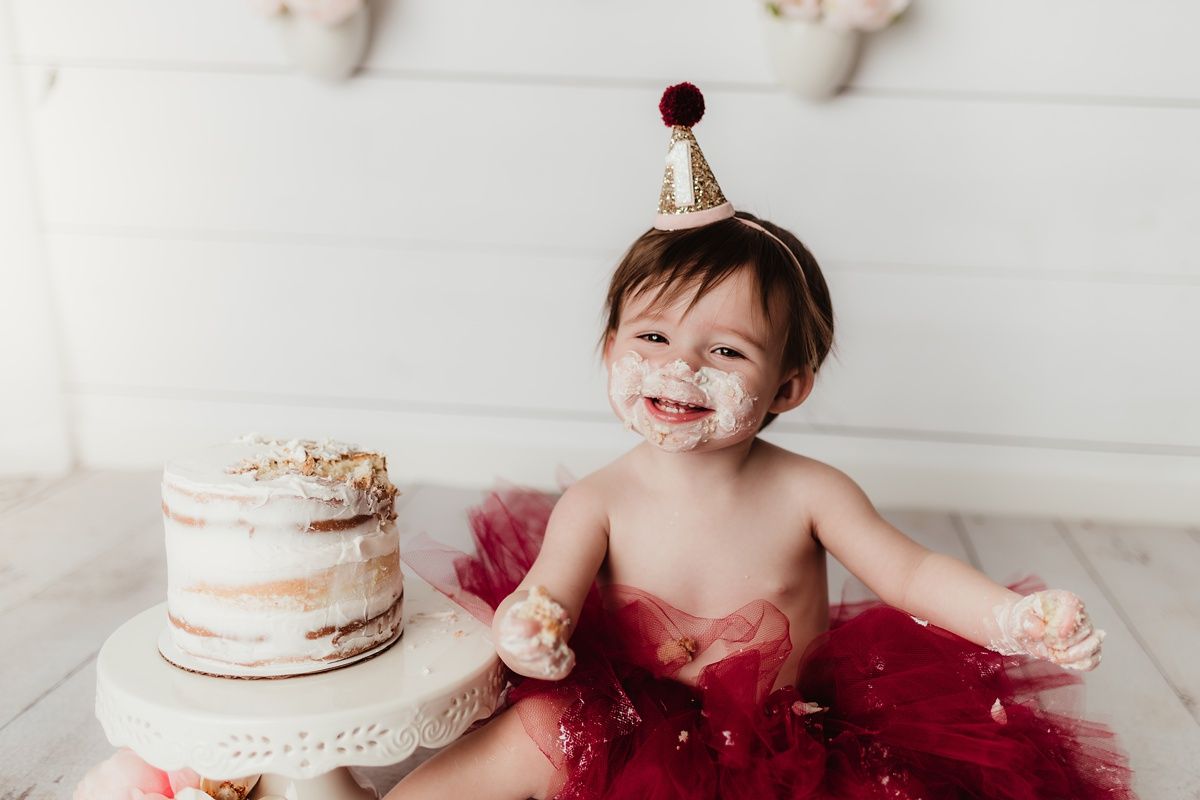 Smiling baby in party hat with messy cake and red tulle skirt.
