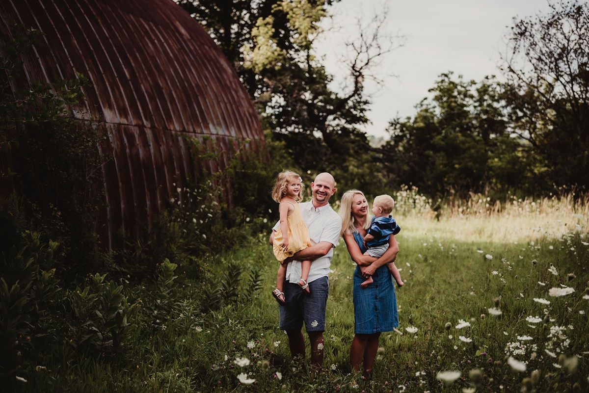 Family stands together near rustic barn in wildflower meadow.