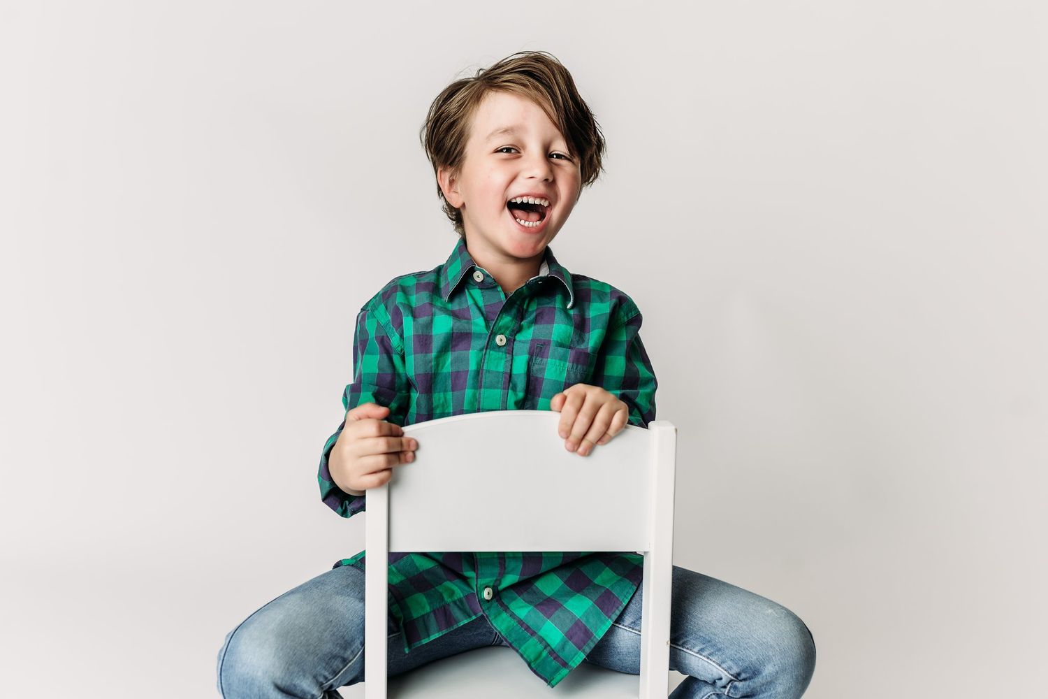 Child laughs while holding onto white chair wearing green plaid shirt and jeans.