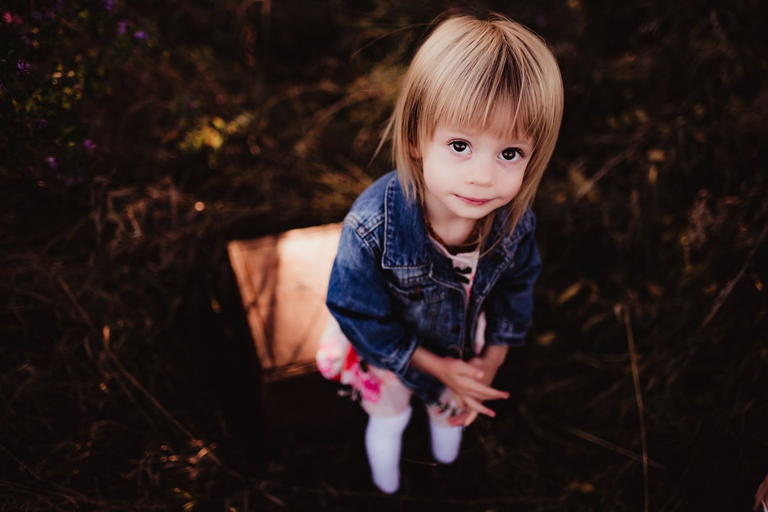 Young child in denim jacket and dress photographed from above in natural setting.