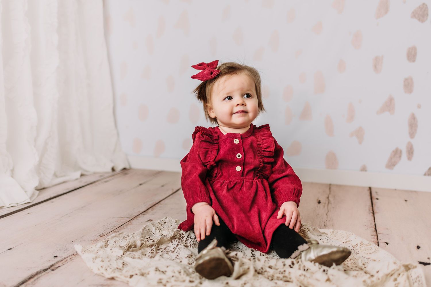 Baby wearing red ruffled dress and bow sitting on wooden floor.