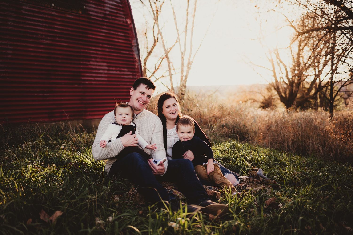 Family relaxing together on grass near a red barn during golden hour.
