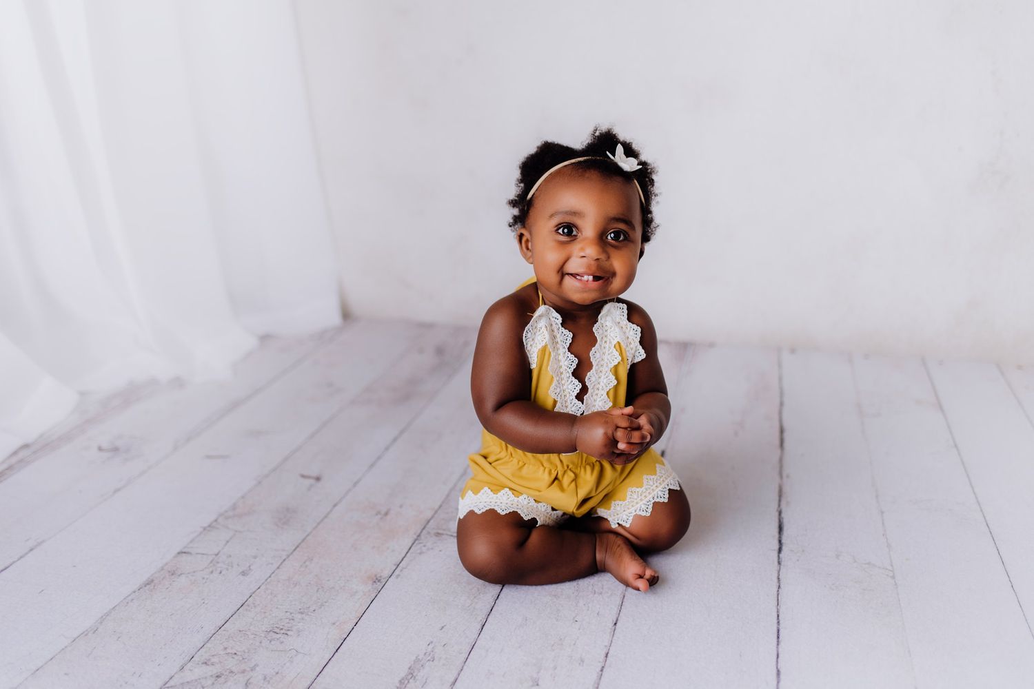 Smiling baby wearing yellow outfit and bow sitting on white floor.