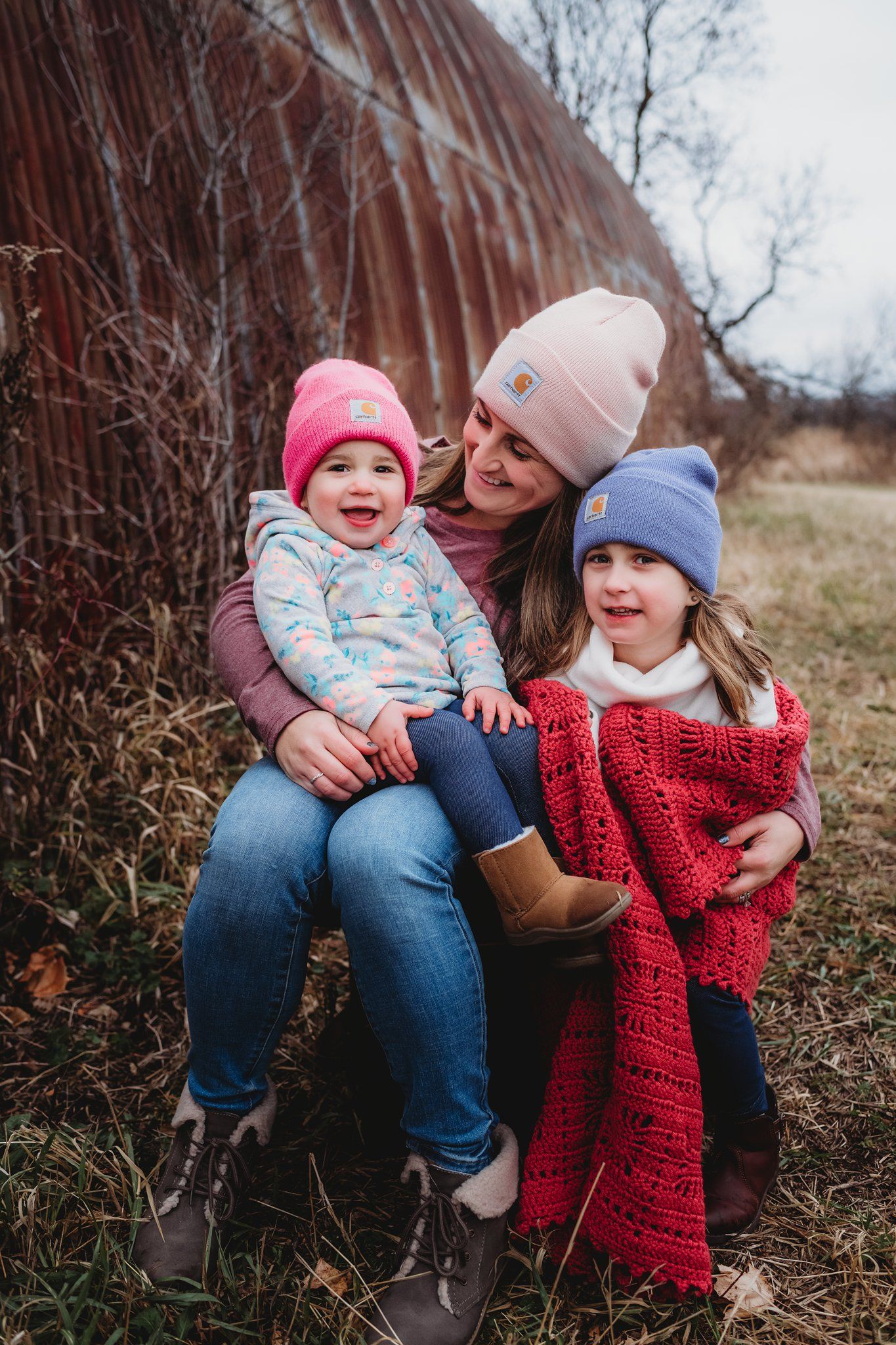 Outdoor family portrait by red barn features cozy winter clothing and bright smiles.