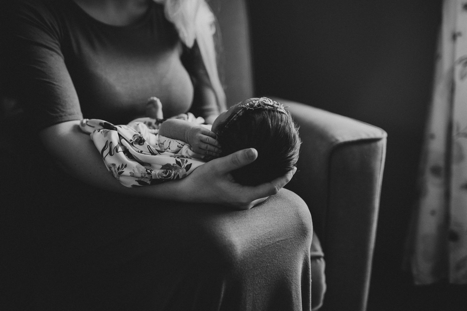 Black and white image of gentle moment with baby being cradled in living room chair.