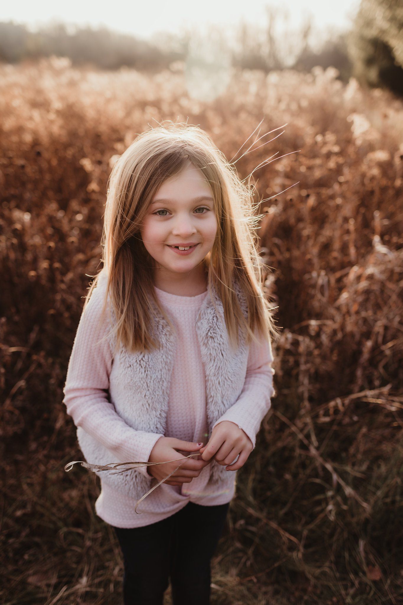 Girl in pink sweater and fur vest smiling in autumn field.