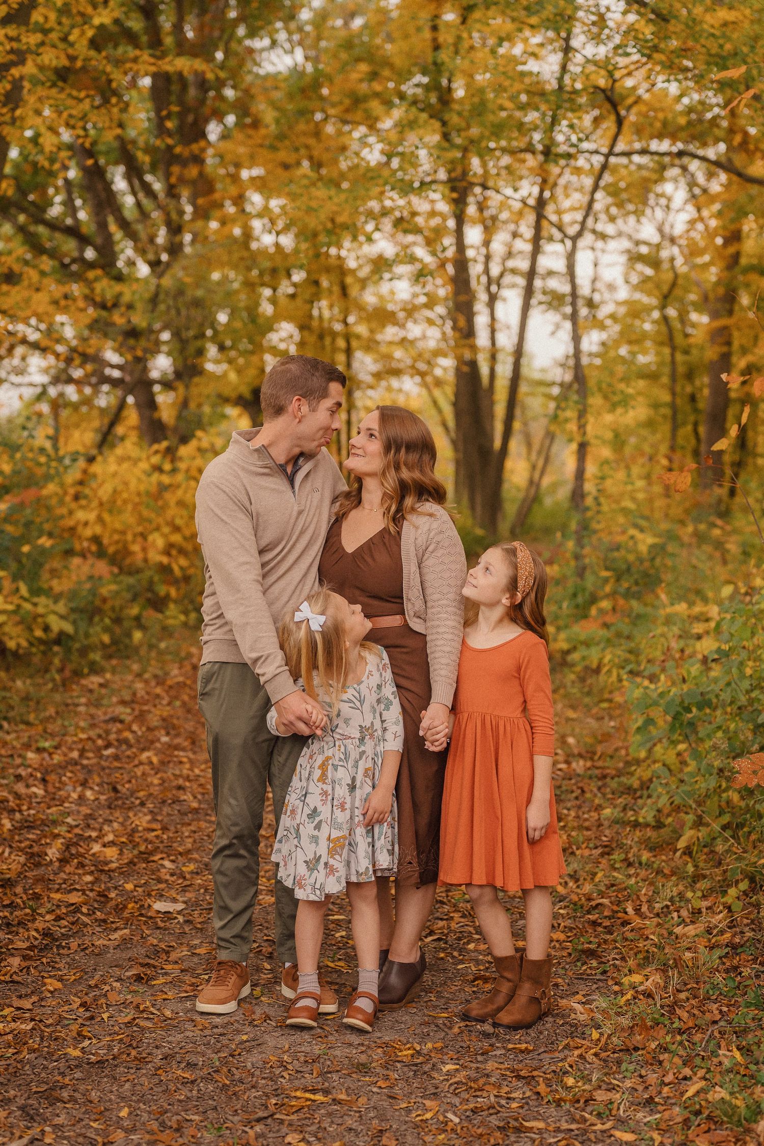 A family in coordinated autumn outfits stands together on a forest path surrounded by golden fall foliage.