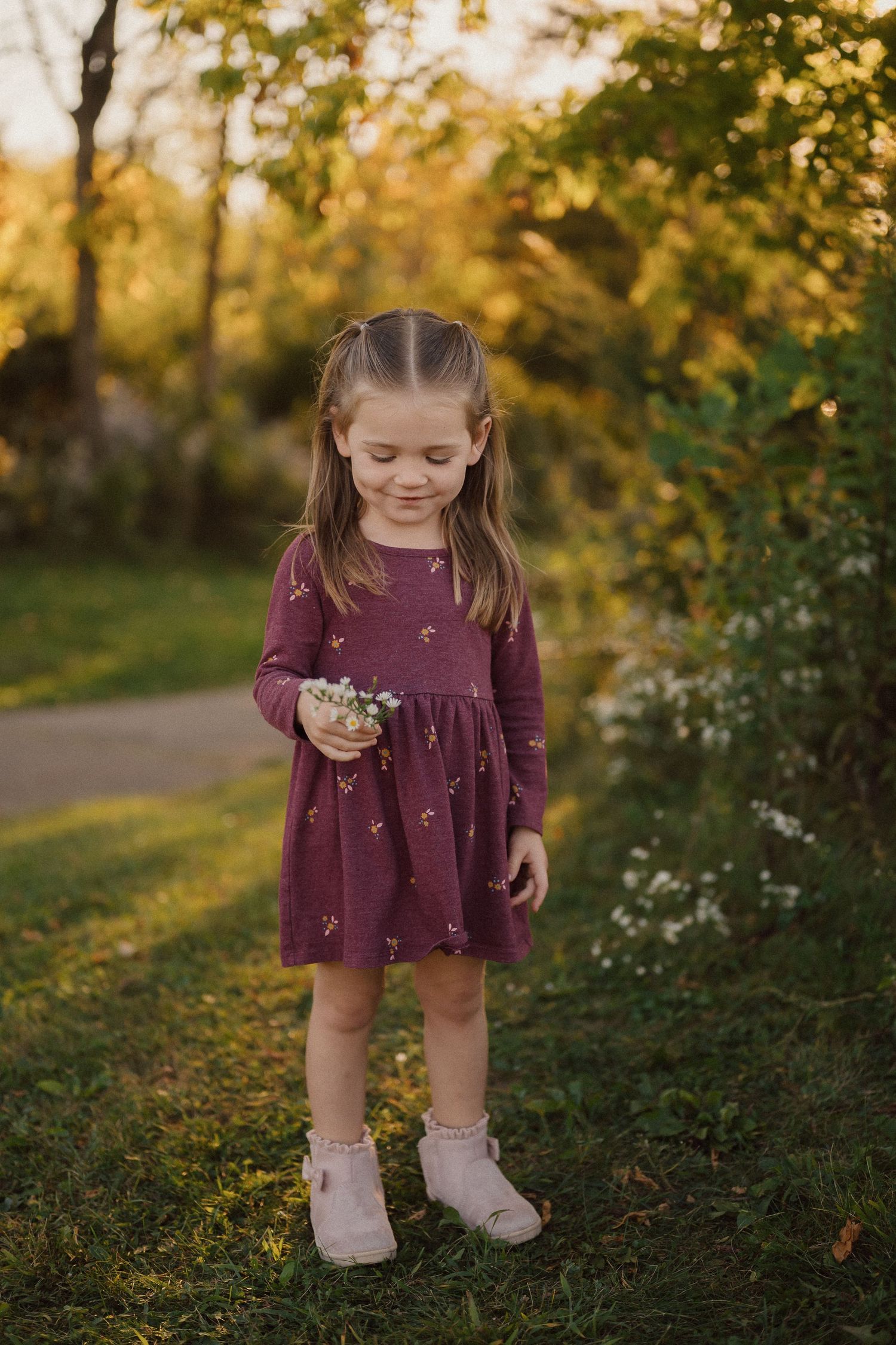Young child in burgundy dress exploring outdoors among wildflowers at sunset.