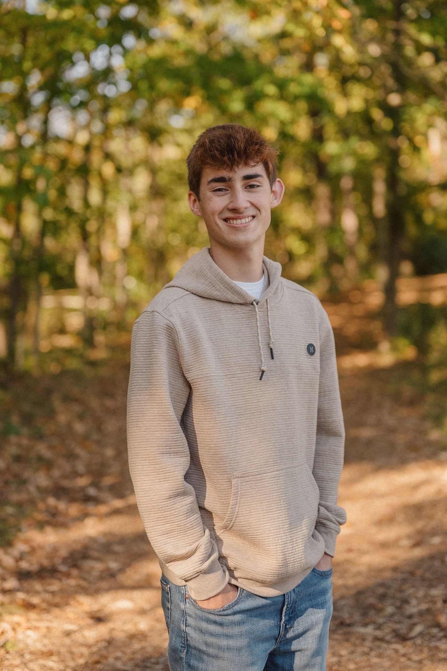Outdoor portrait against fall foliage wearing a light gray hoodie and jeans.