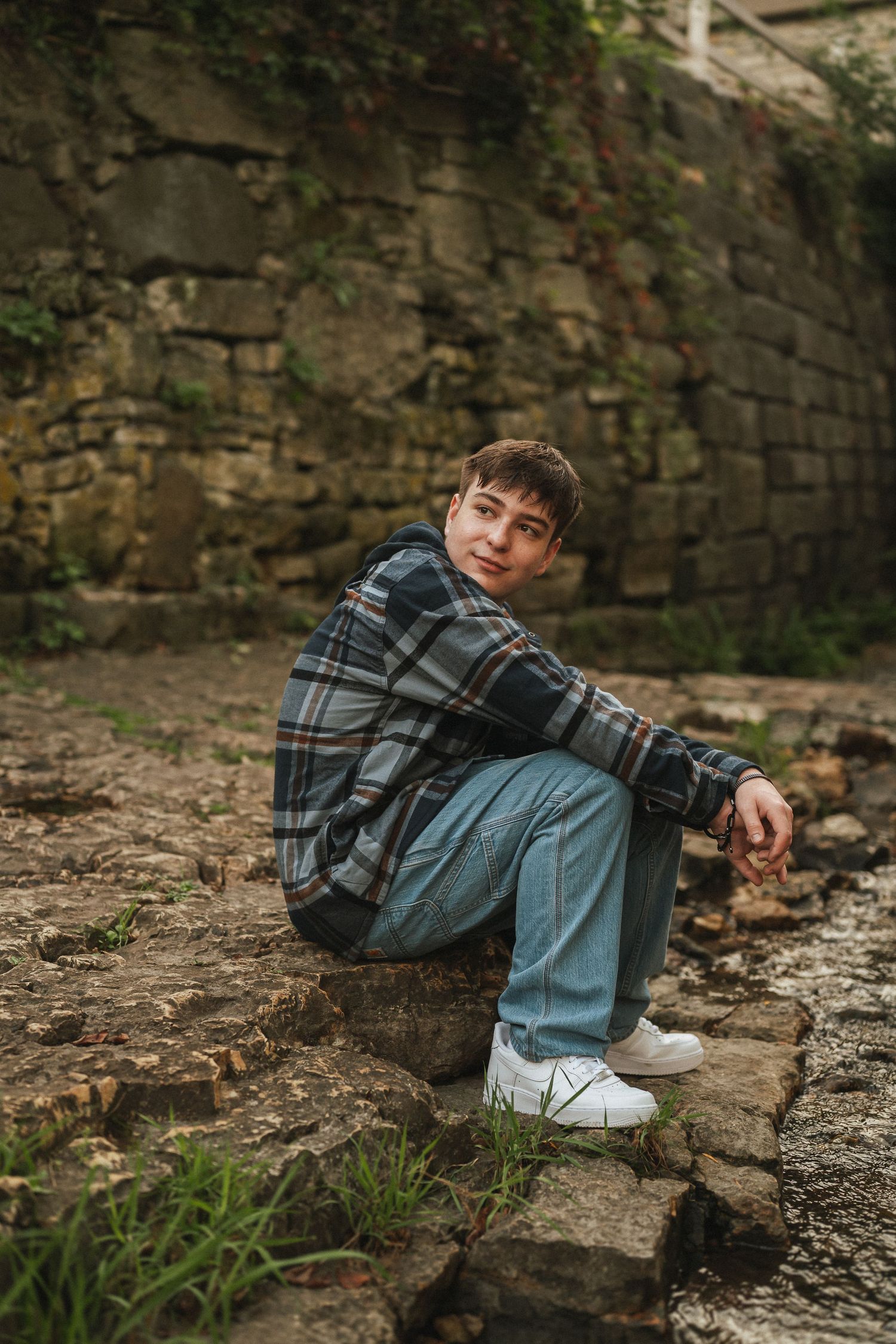 A person in a plaid jacket and jeans sits casually on stone steps against an old brick wall.