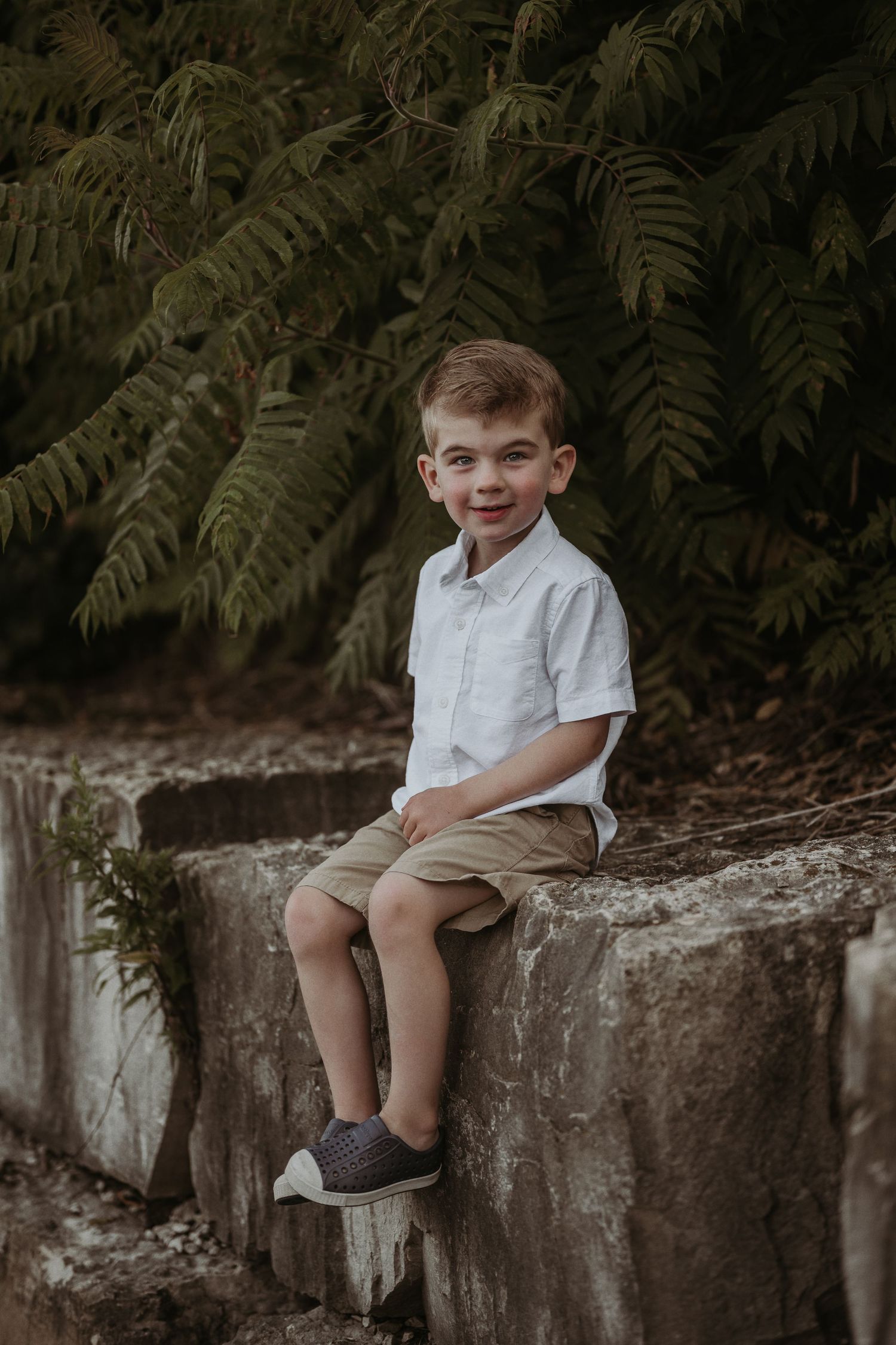 A cheerful child in white shirt sits on stone wall with ferns in background.