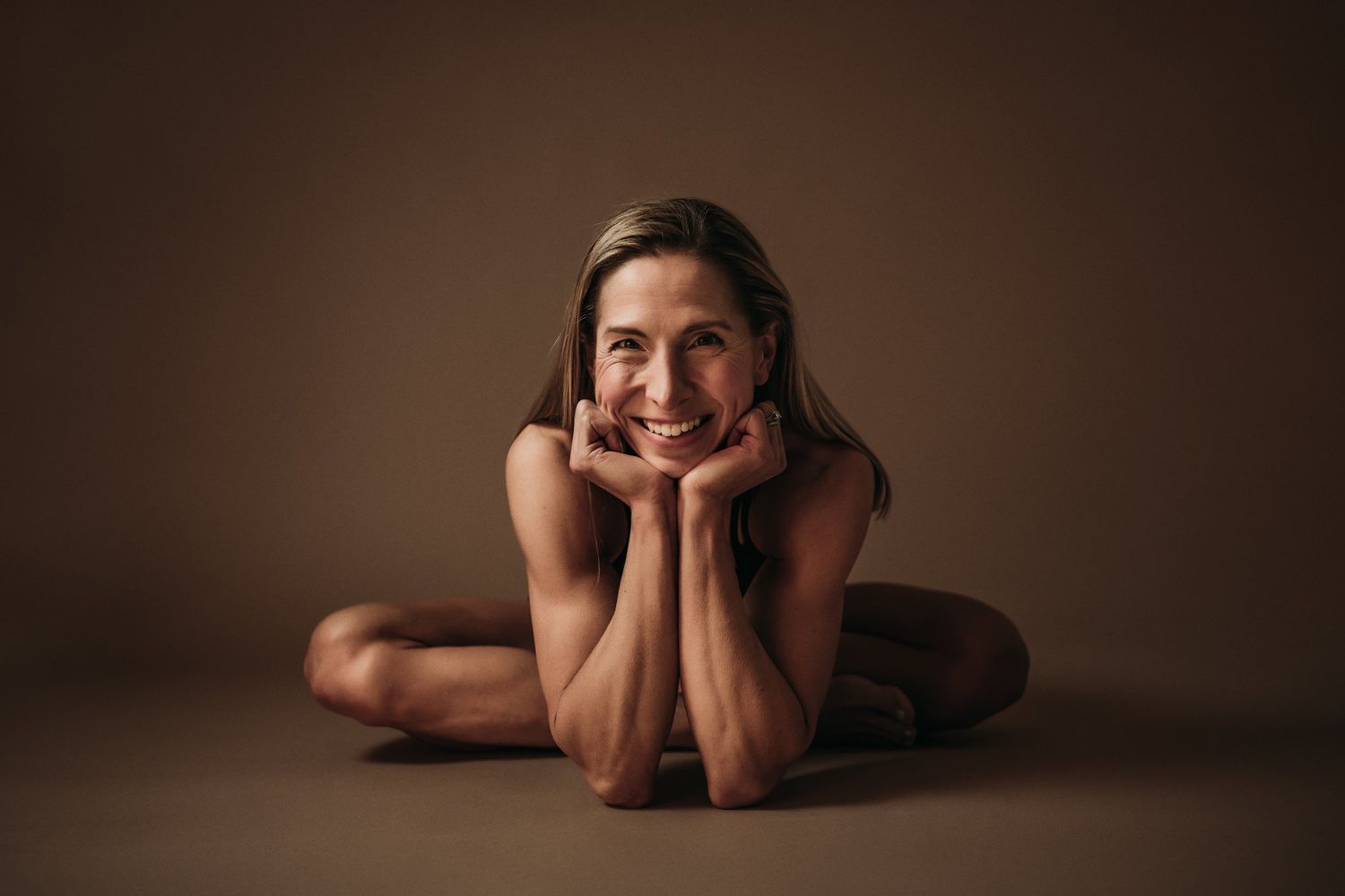 Joyful fitness model in relaxed pose on brown studio floor.